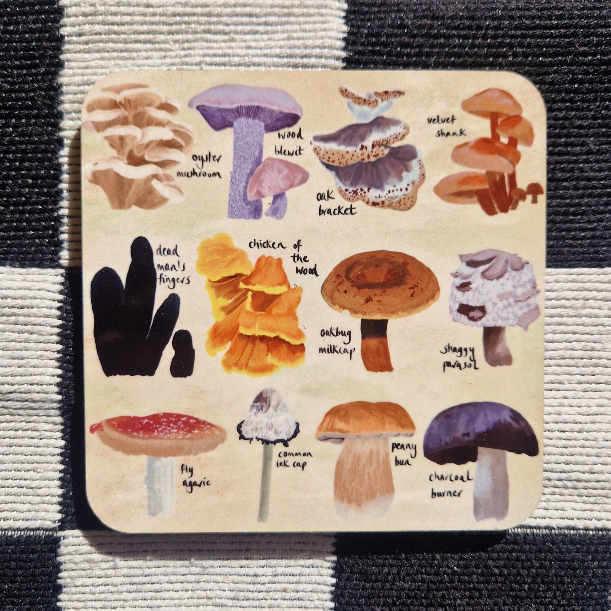 A square coaster featuring 12 different fungi illustrations on a cream background. Behind the coaster is a black and white checkered fabric background.