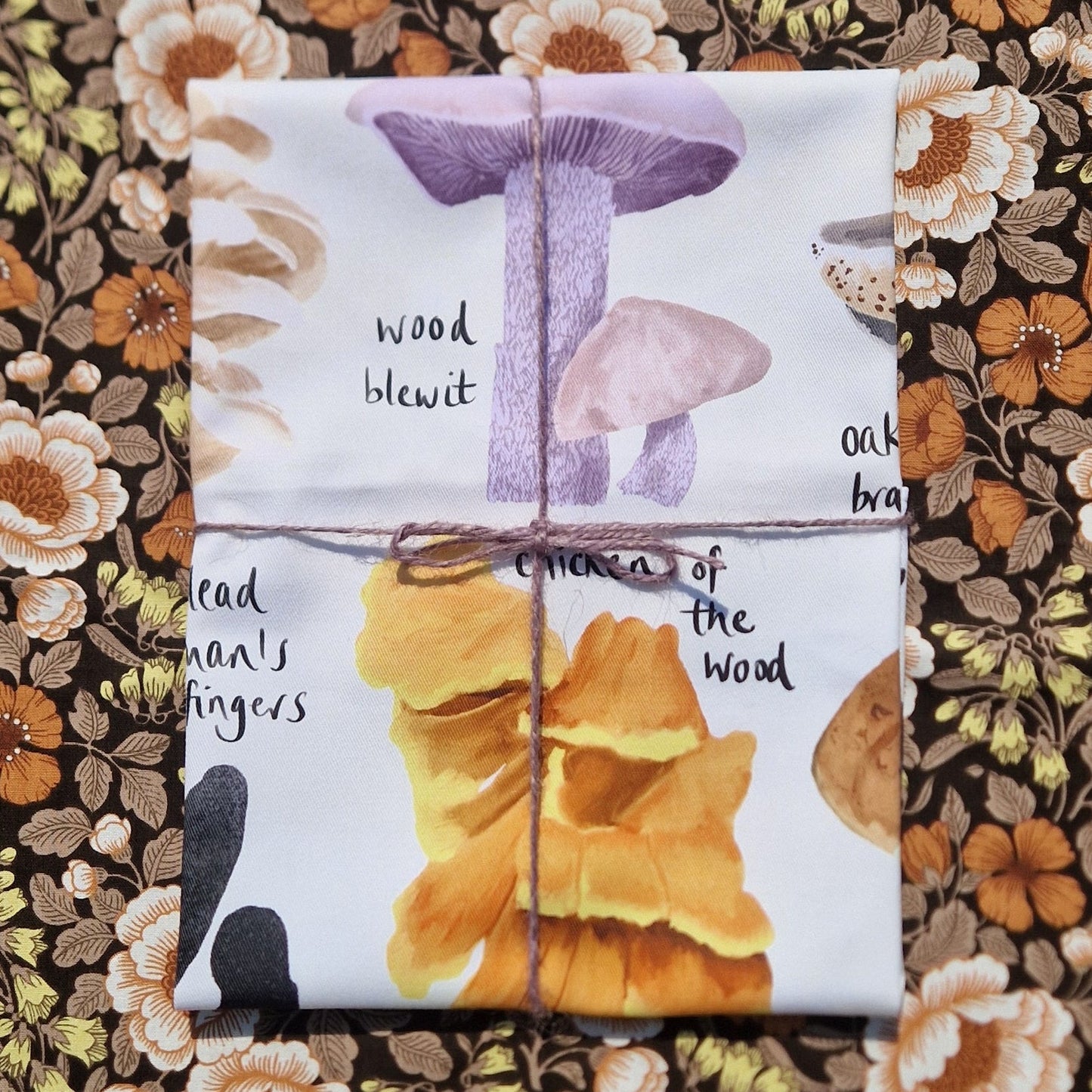 A folded white teatowel with fungi illustrations on it wrapped it purple macrame twine sits on a vintage floral patterned fabric.