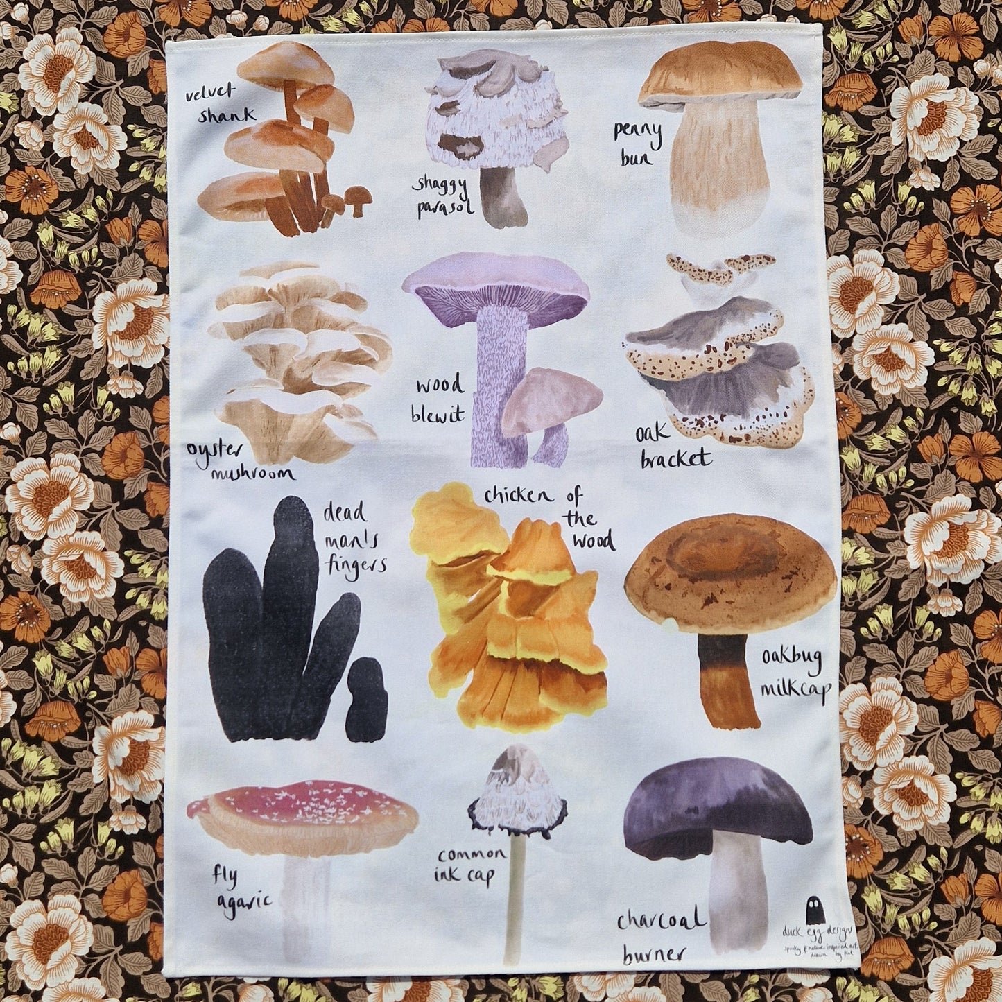 A white tea towel sits on a retro floral printed fabric. The tea towel features 12 different full colour fungi illustrations with their names next to them in black handwriting. 