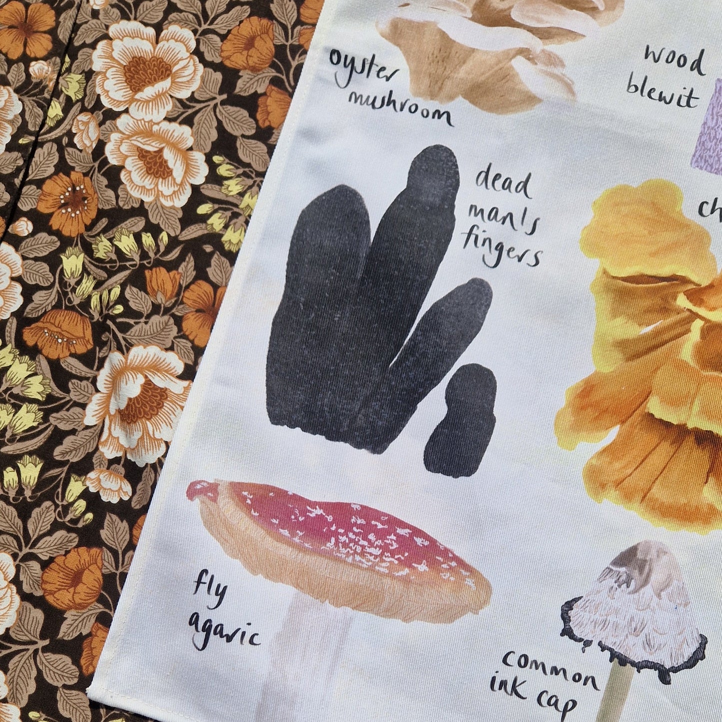 The bottom left corner of a white tea towel with colourful fungi illustrations sits on a retro floral fabric.