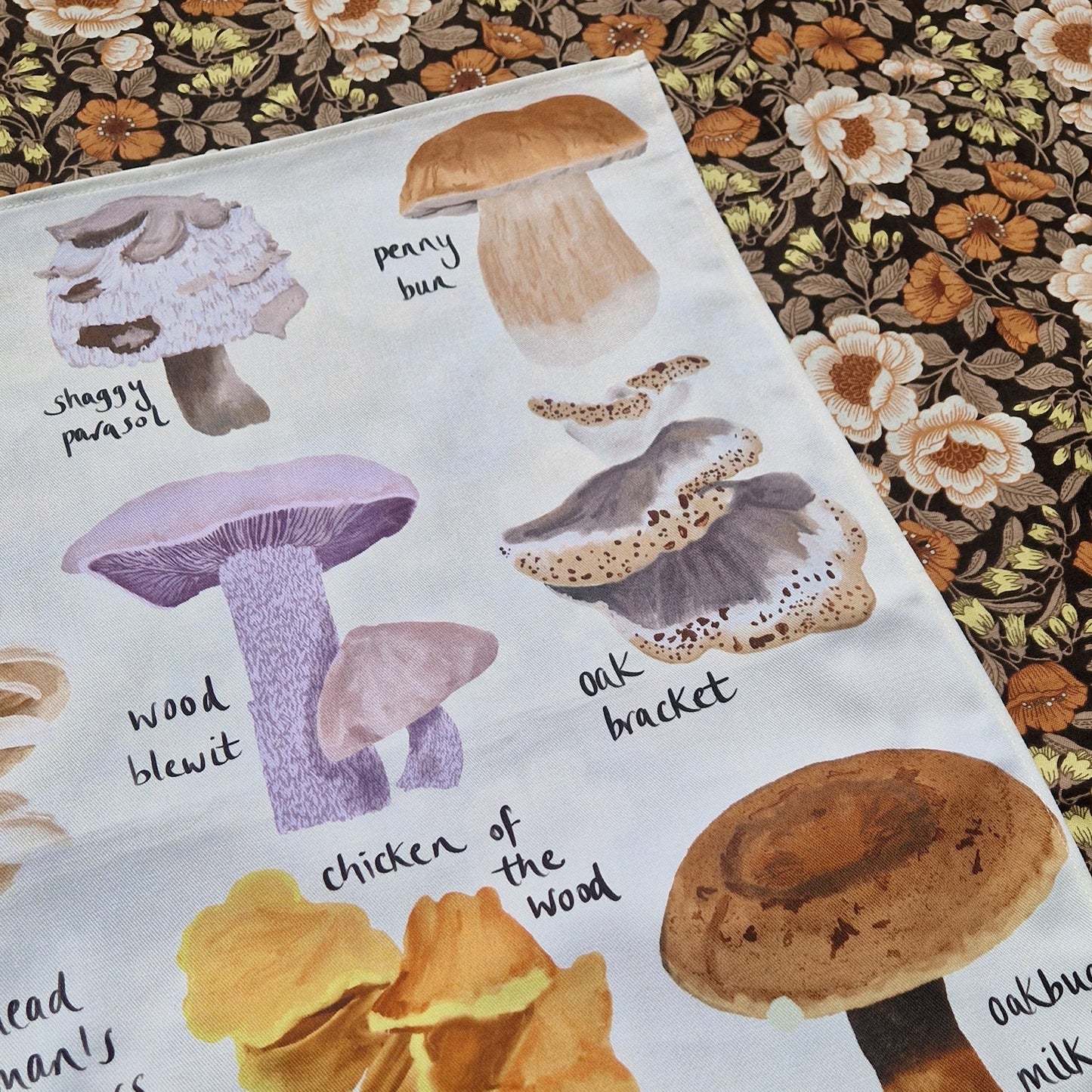 A white tea towel featuring illustrations of different fungi sits on a retro floral fabric.