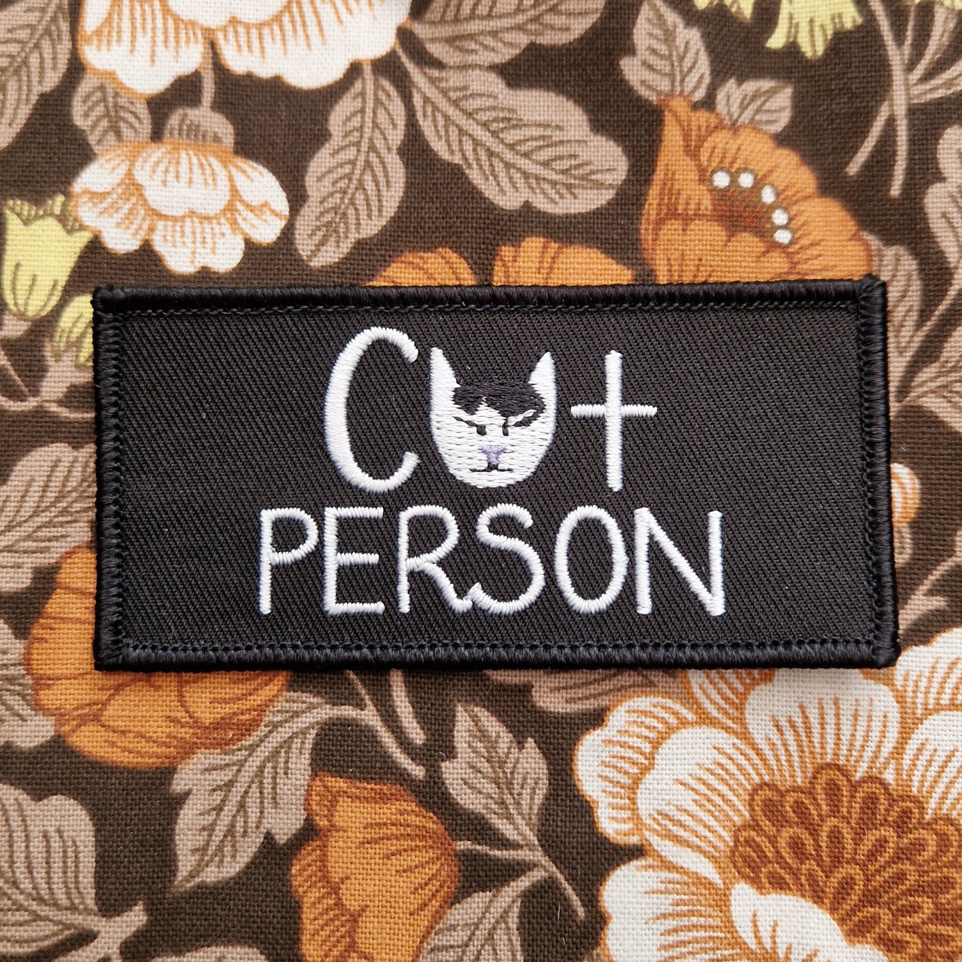 A black rectangular embroidered patch with the words 'Cat Person' in white with a black and white cat head where the a in cat should be. Behind the patch is a floral patterned fabric.