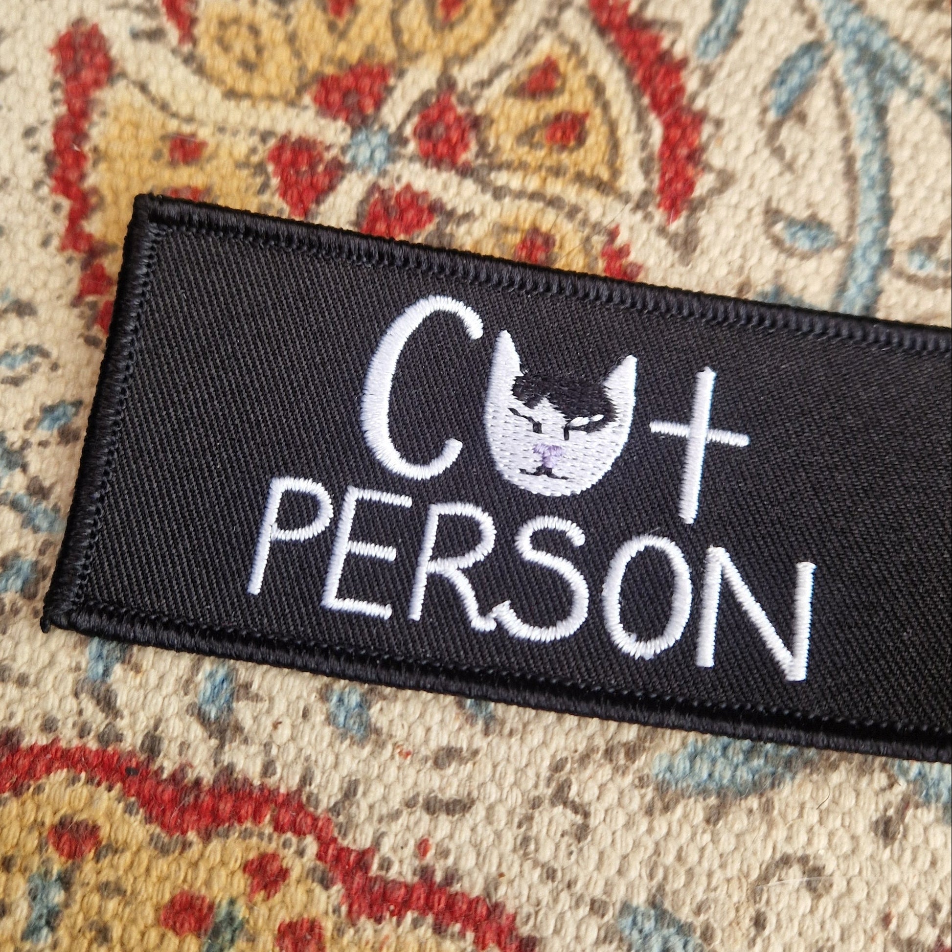  A close up of a black rectangular embroidered patch with the words 'Cat Person' in white with a black and white cat head where the a in cat should be. Behind the patch is a patterned fabric.