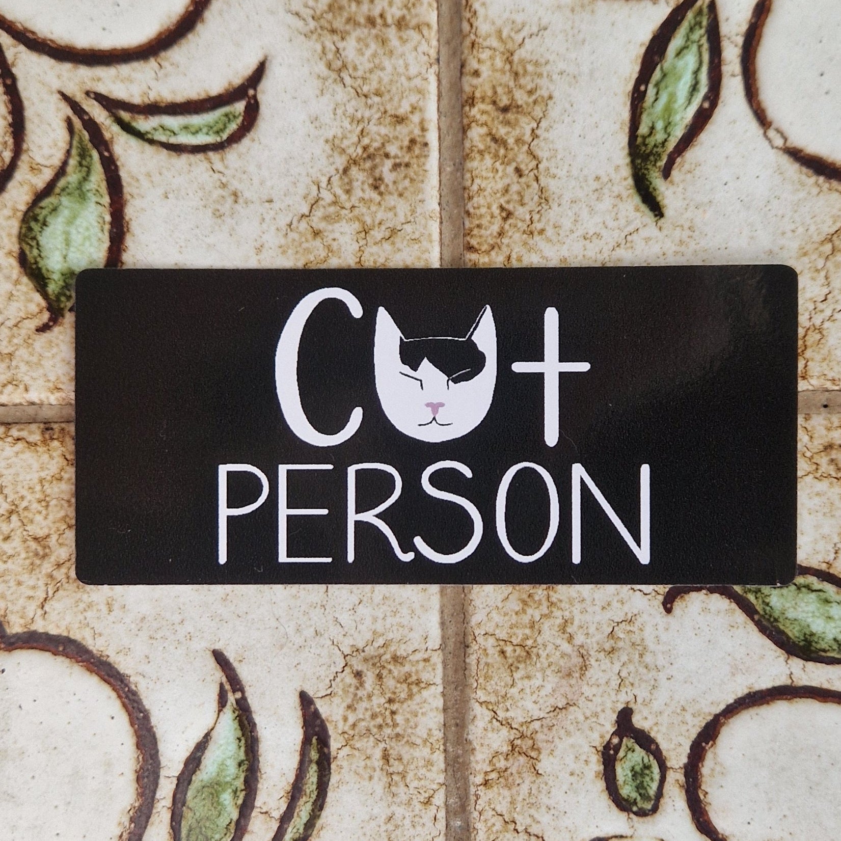 A black rectangular sticker featuring the words 'CAT PERSON' across the middle in white handwriting with a white and black cat head instead of an A. The sticker sits on a patterned tile background.