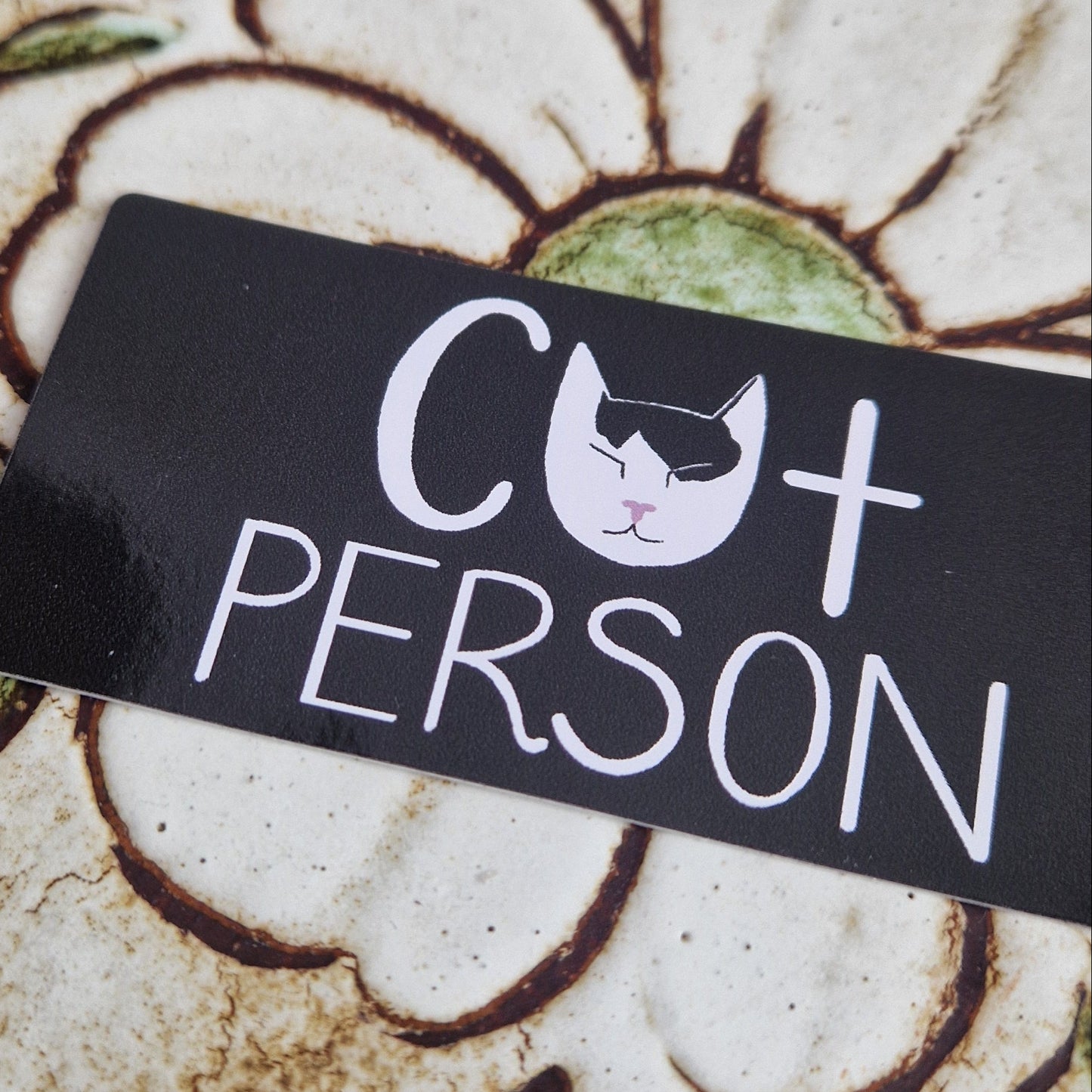 A closer look at the cat person vinyl sticker showing the detail in the cat head which is in the place of an A. The sticker sits on a floral tiled background.