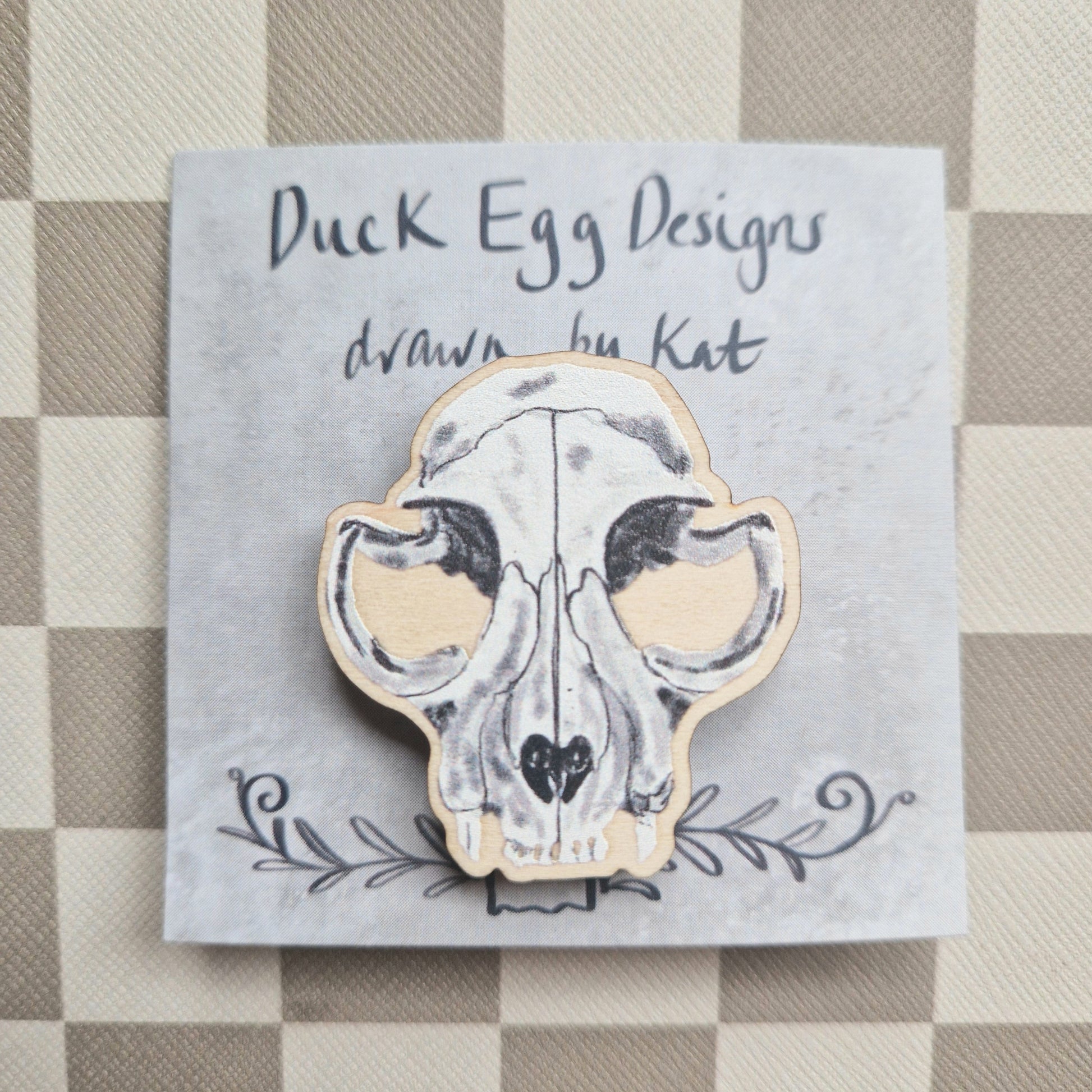 A white cat skull pin badge sits on a grey backing card in front of a checkered background.