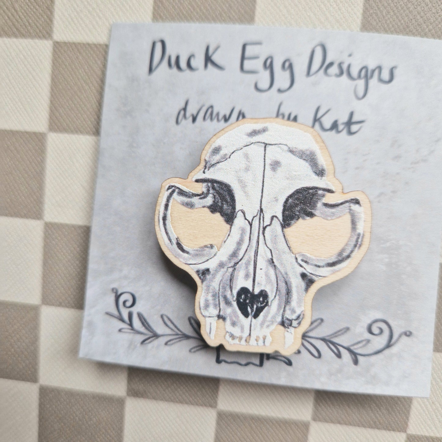 A white, grey and black cat skull pin badge sits on a grey backing card with the words 'Duck Egg Designs drawn by Kat' across the top. Behind the backing card you can see a checkered cream and brown background.