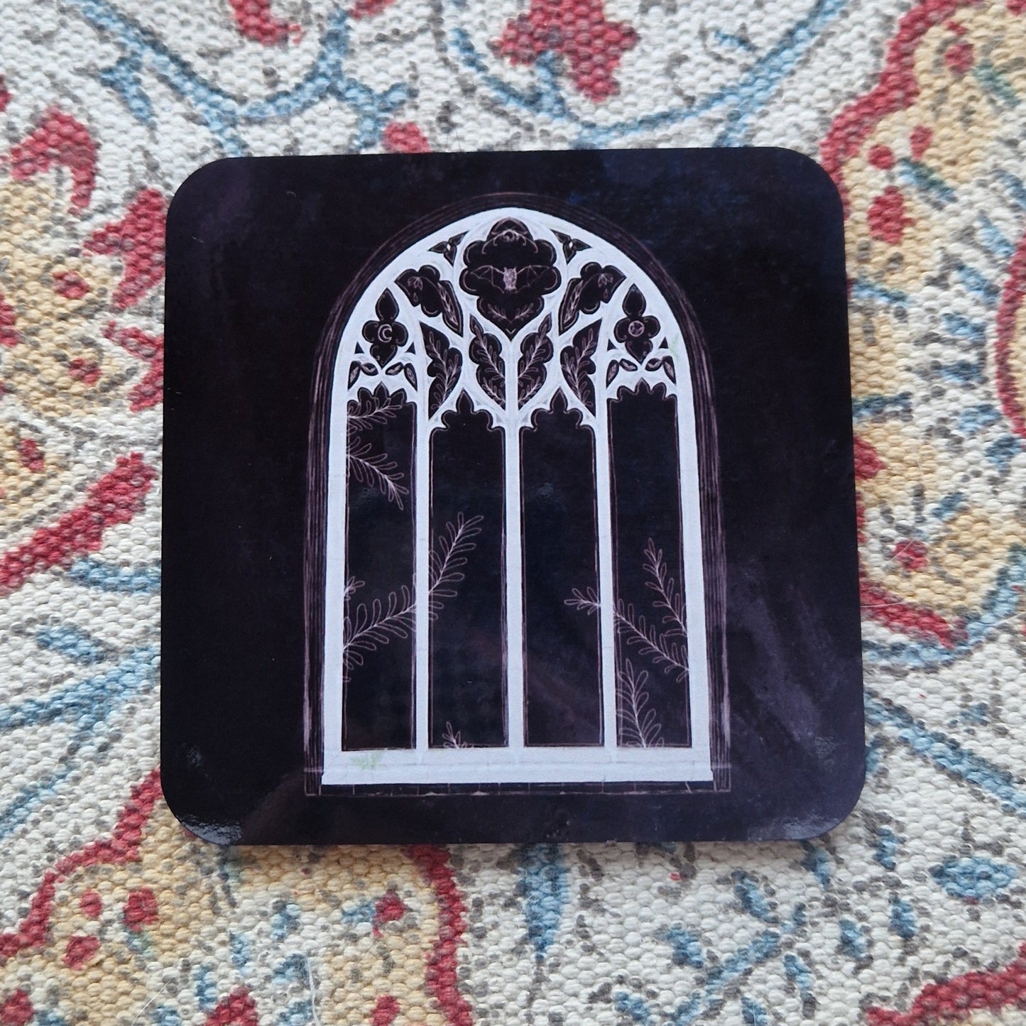 A black square coaster with a cathedral window design sits on a patterned fabric background.