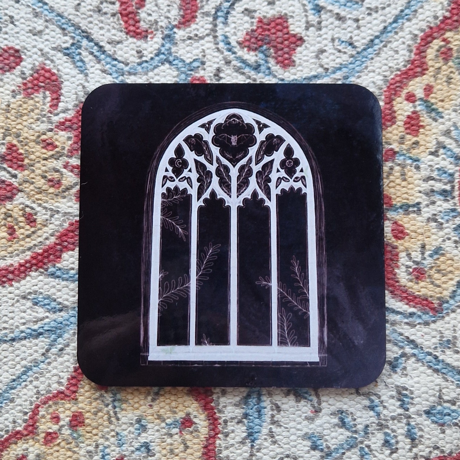 A black square coaster with a cathedral window design sits on a patterned fabric background.