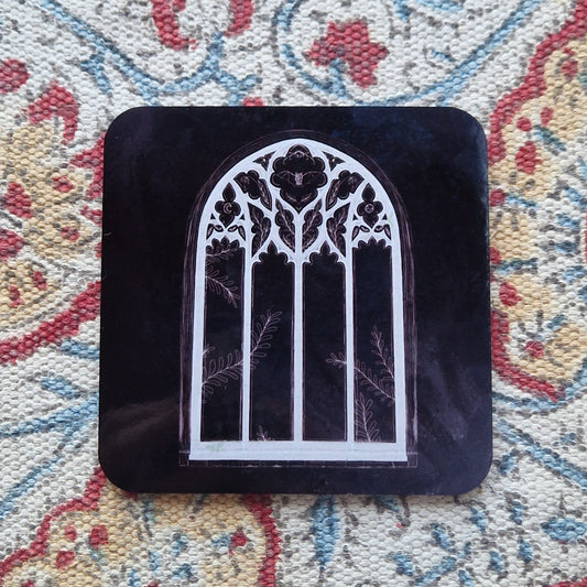 A black square coaster with a cathedral window design sits on a patterned fabric background.