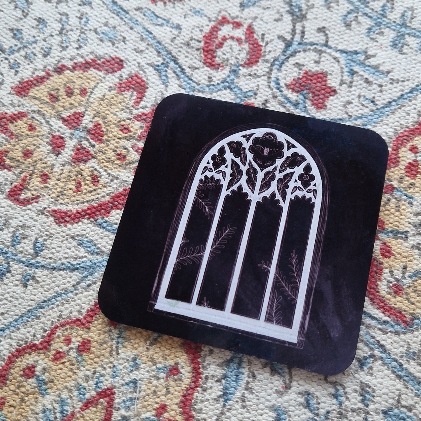 A black square coaster with a cathedral window design sits on a patterned fabric background.