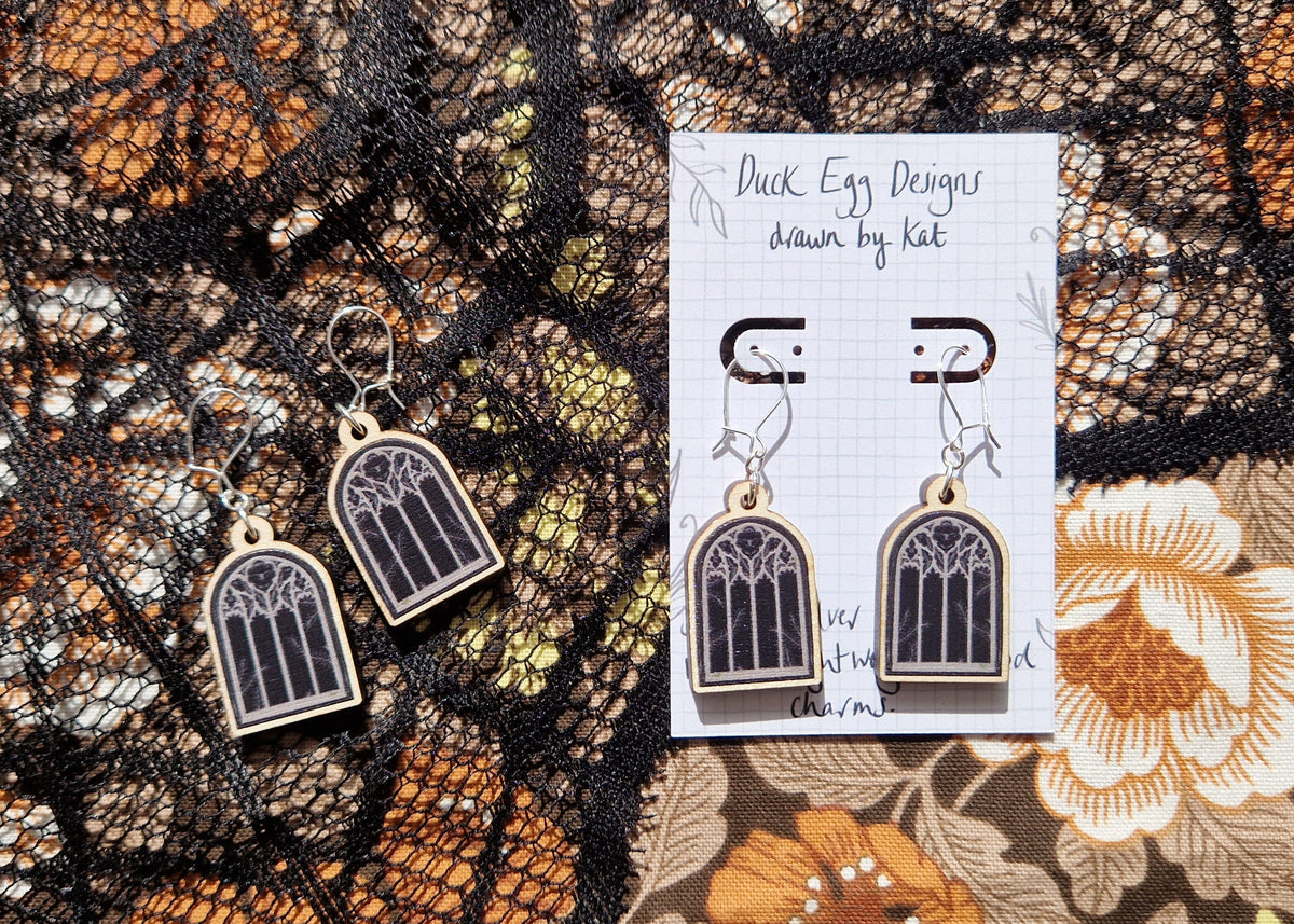 Two pairs of black cathedral window earring sit next to each other on a floral fabric and black lace background. The pair of earrings on the right sit on a white backing card with the words 'Duck Egg Designs drawn by Kat' in black handwriting.