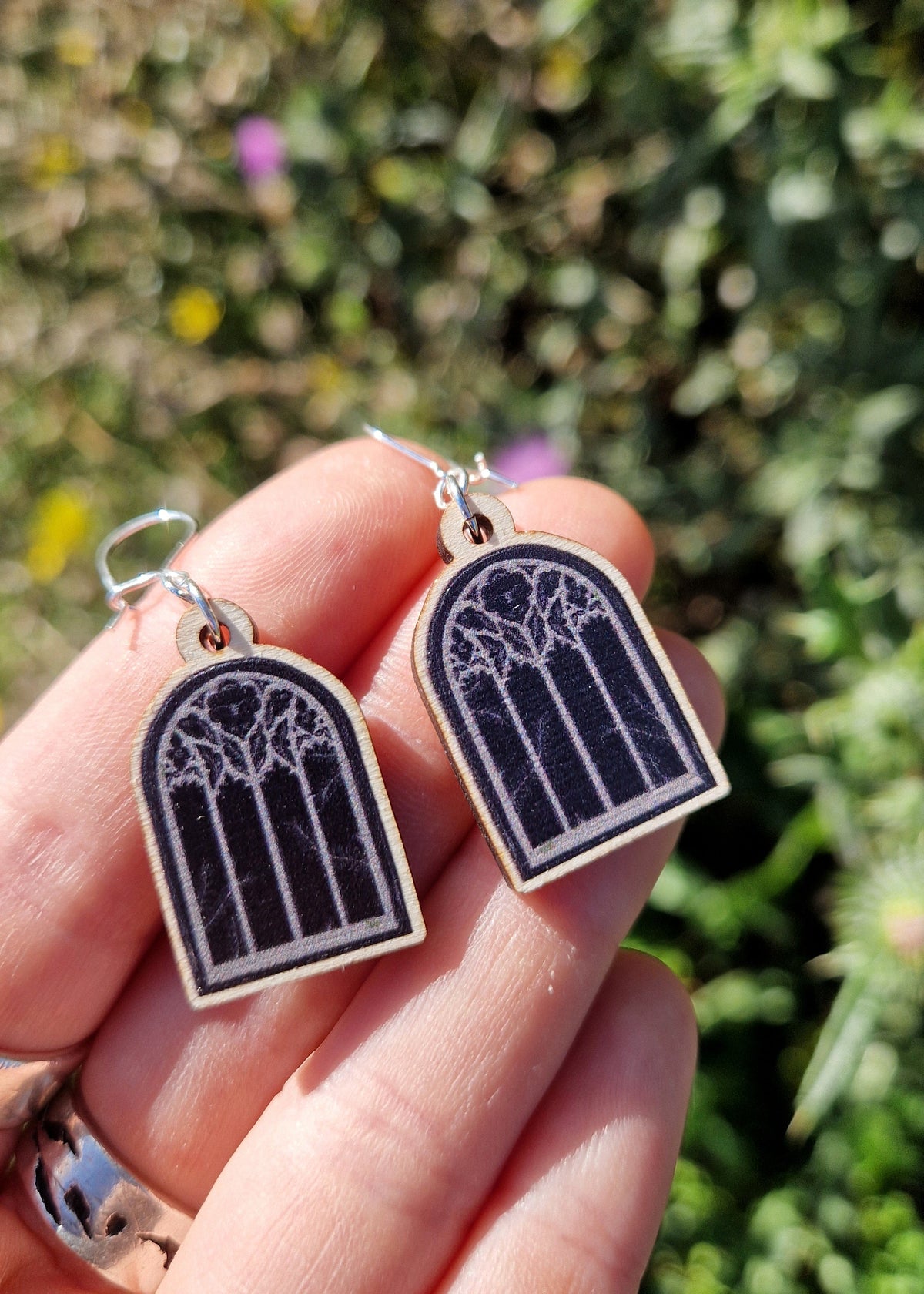 A pair of black cathedral window earrings with white and silver details sit on a white hand over some out of focus green flower plants.