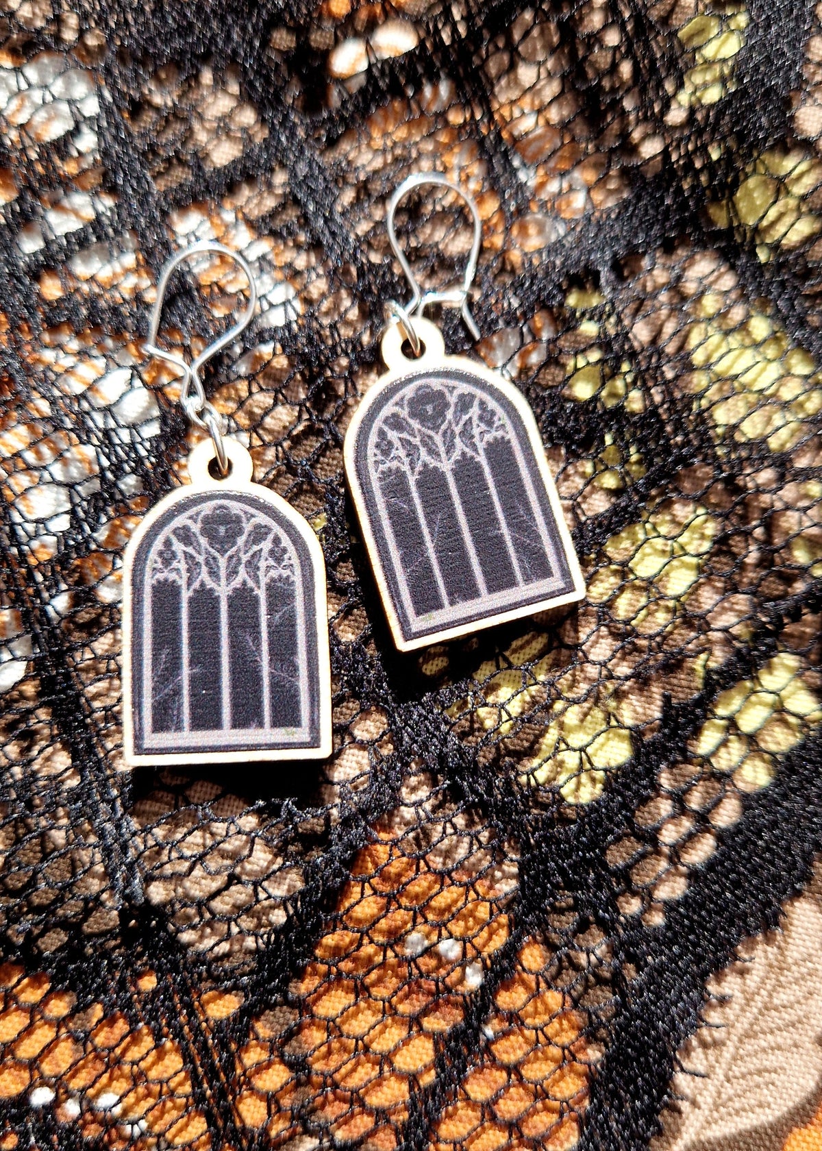 A pair of gothic window earrings sit on a black lace and floral fabric background.