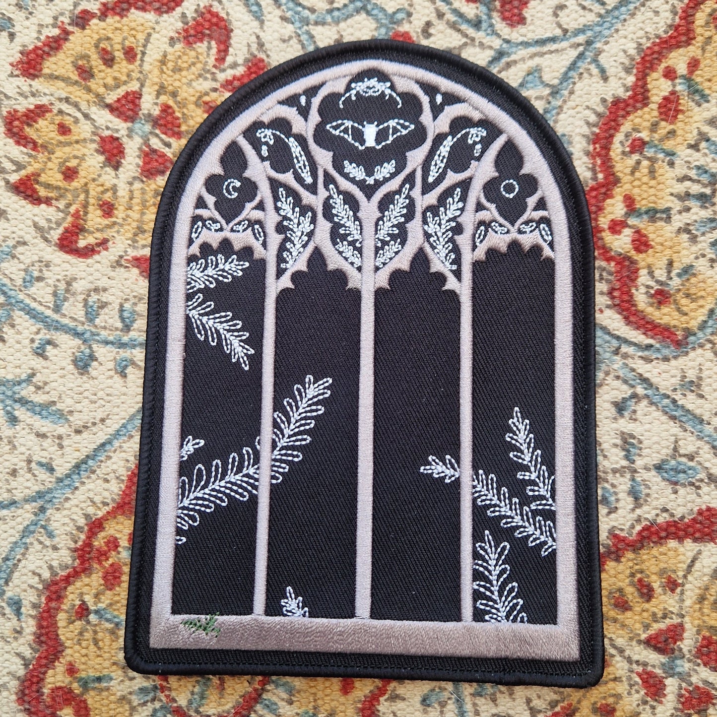 A black arched window shaped patch sits on a patterned fabric background. The patch has a gothic window design with a fern growing in the bottom left of the window and a bat and nature inspired design in the top section of the window.