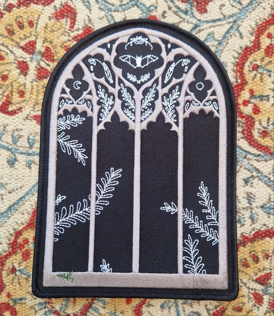 A black arched window shaped patch sits on a patterned fabric background. The patch has a gothic window design with a fern growing in the bottom left of the window and a bat and nature inspired design in the top section of the window.