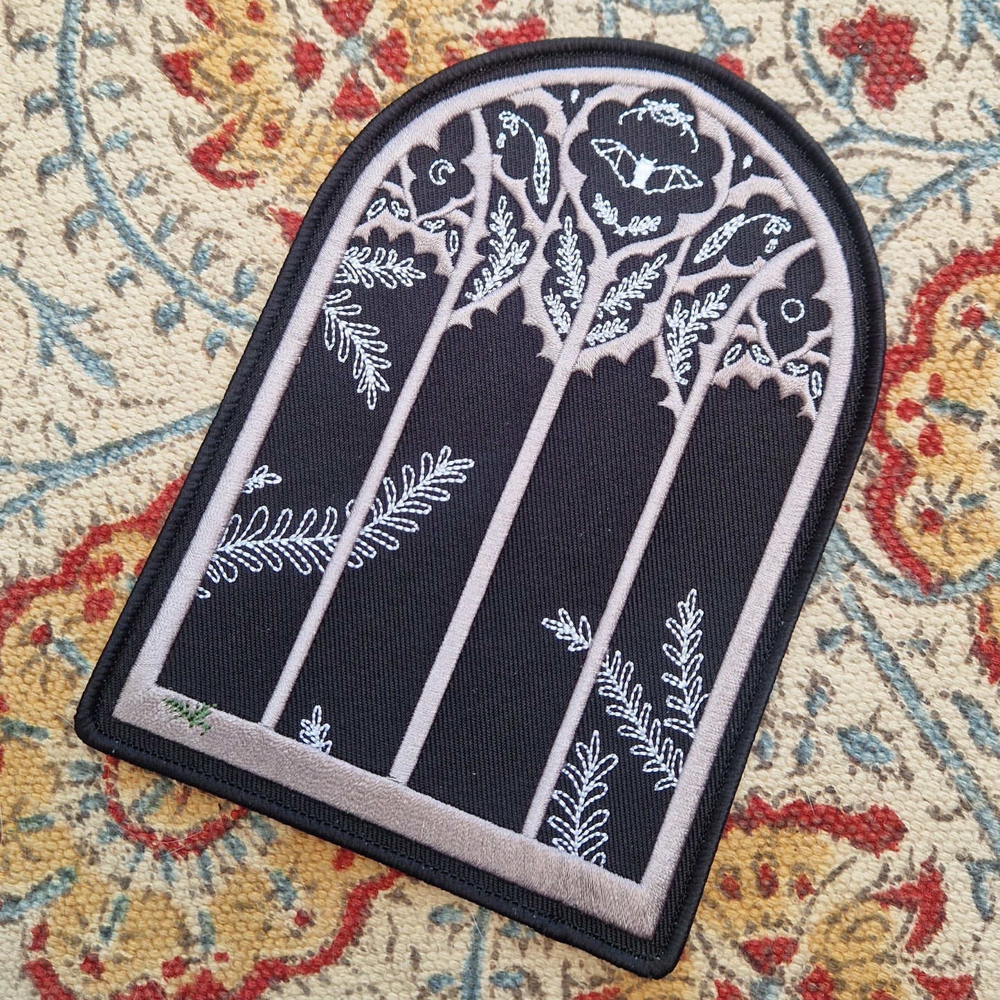 A black arched window shaped patch sits on a patterned fabric background. The patch has a gothic window design with a fern growing in the bottom left of the window and a bat and nature inspired design in the top section of the window.