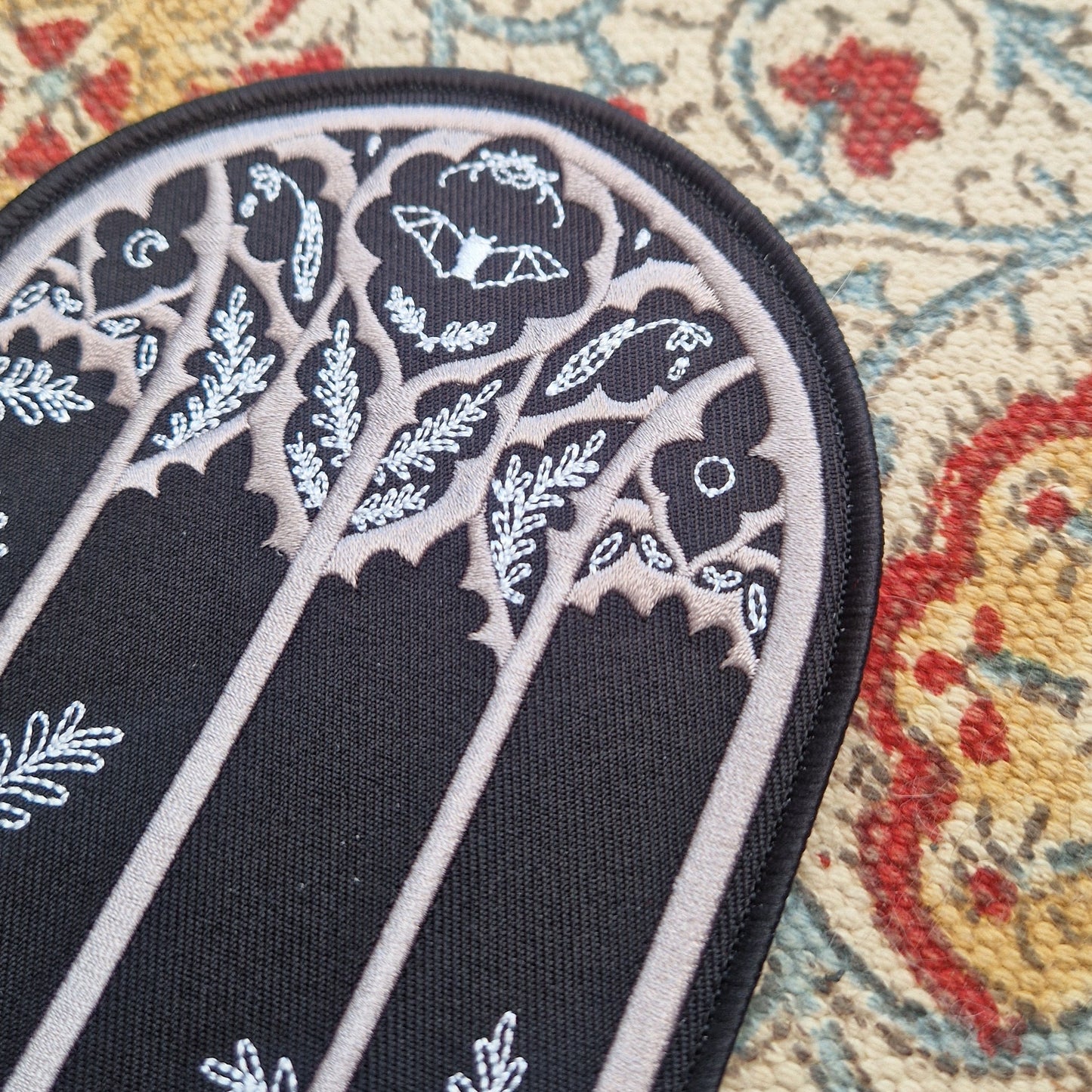 A close up of the gothic cathedral window inspired patch showing the nature filled window design featuring a bat, leafy fronds, flowers and the moon.
