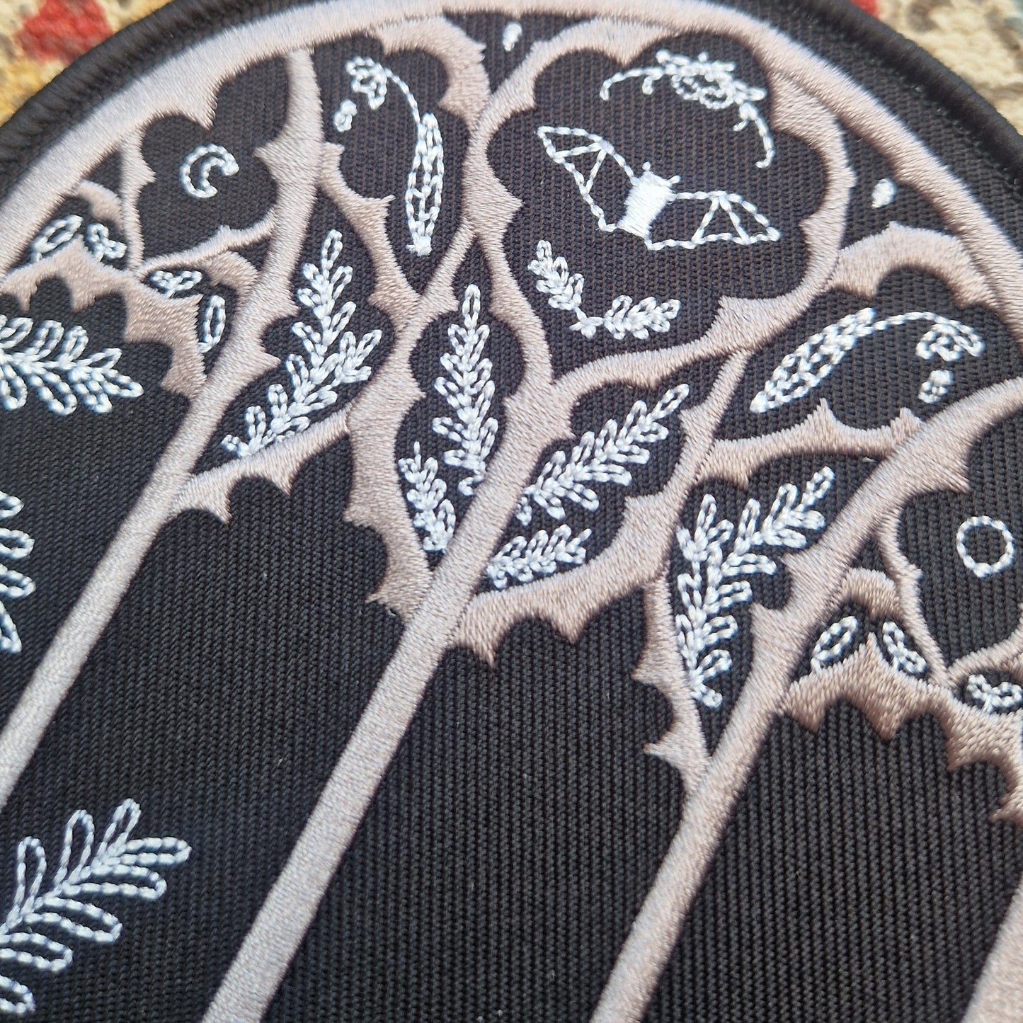 A close up of the gothic cathedral window inspired patch showing the nature filled window design featuring a bat, leafy fronds, flowers and the moon.
