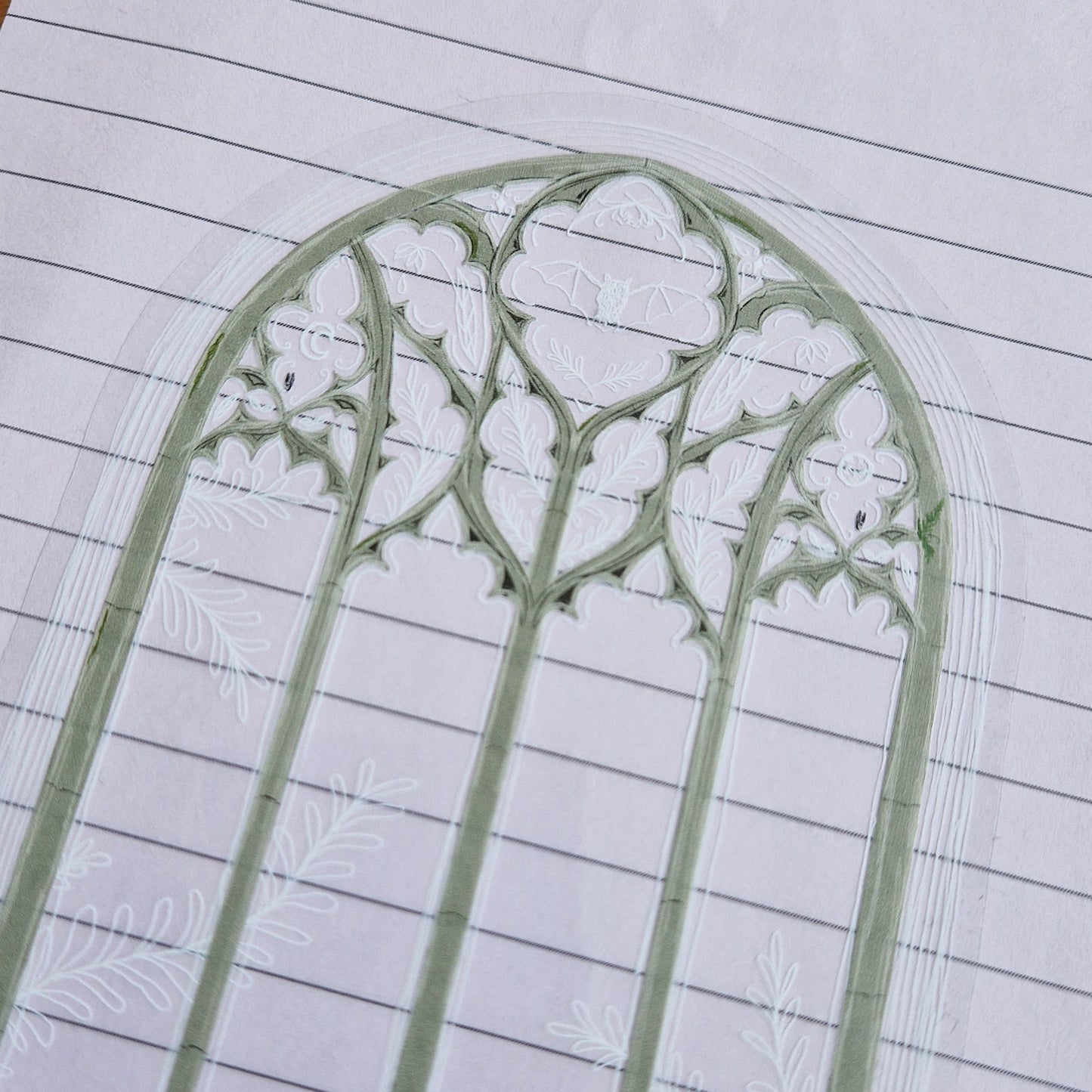 A close up view of the top of the sticker showing the detail in the cathedral window illustration. The outline of the window is in grey brick, with white line sketch details. Inside the top panes of glass are a bat illustration, flowers, two moons and leafy frond designs. On the top right of the window frame there is a small green fern plant. The sticker has a transparent background and is stuck to a lilac lined sheet of paper.