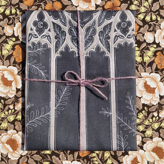 A dark grey teatowel sits on a retro floral patterned background. The teatowel is wrapped in purple twine and you can see part of a gothic cathedral window design.