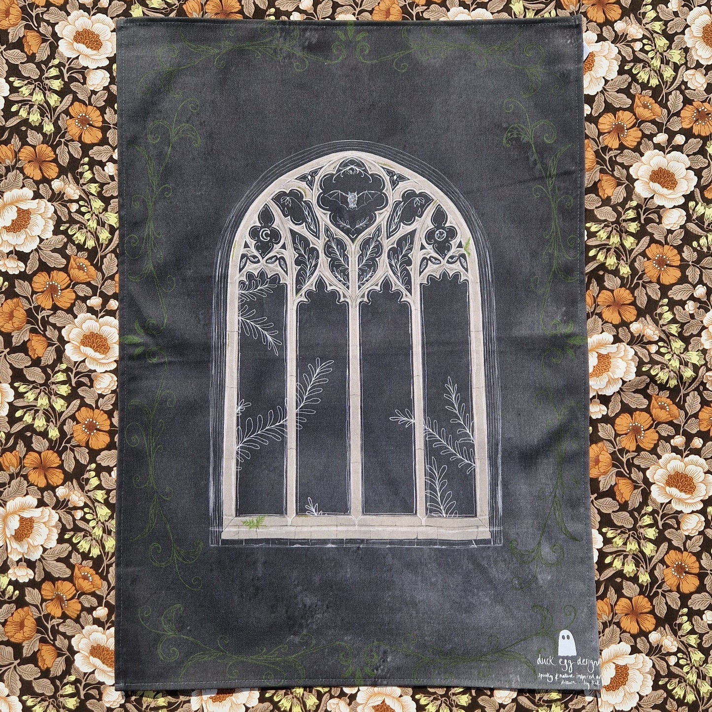 A dark grey tea towel sits on a retro floral patterned fabric. In the middle of the teatowel is a cathedral window design with a bat, leafy plant and moon design in it. Around the edges of the teatowel is a green leafy vine design and on the bottom right corner is a white Duck Egg Designs logo.