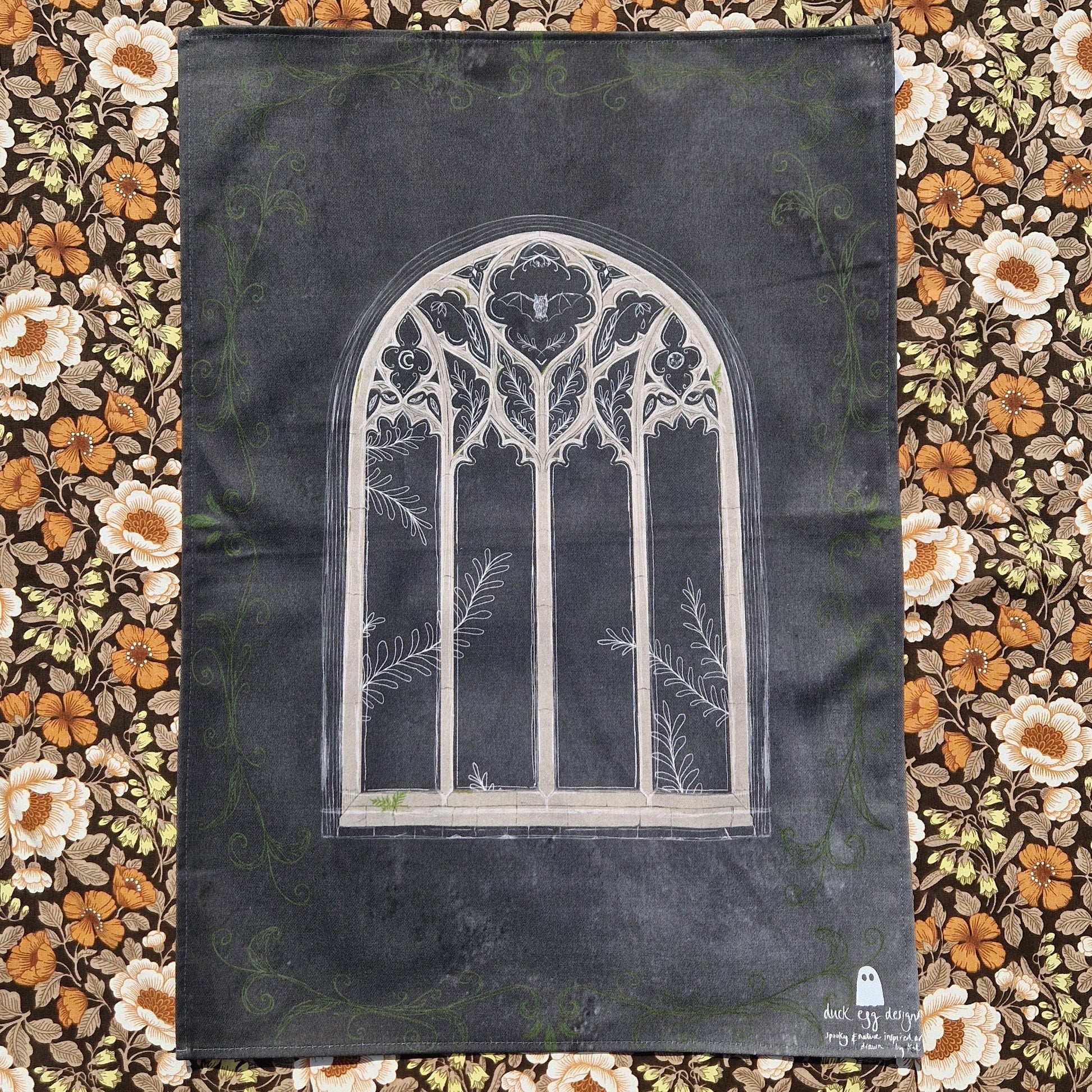 A dark grey tea towel sits on a retro floral patterned fabric. In the middle of the teatowel is a cathedral window design with a bat, leafy plant and moon design in it. Around the edges of the teatowel is a green leafy vine design and on the bottom right corner is a white Duck Egg Designs logo.