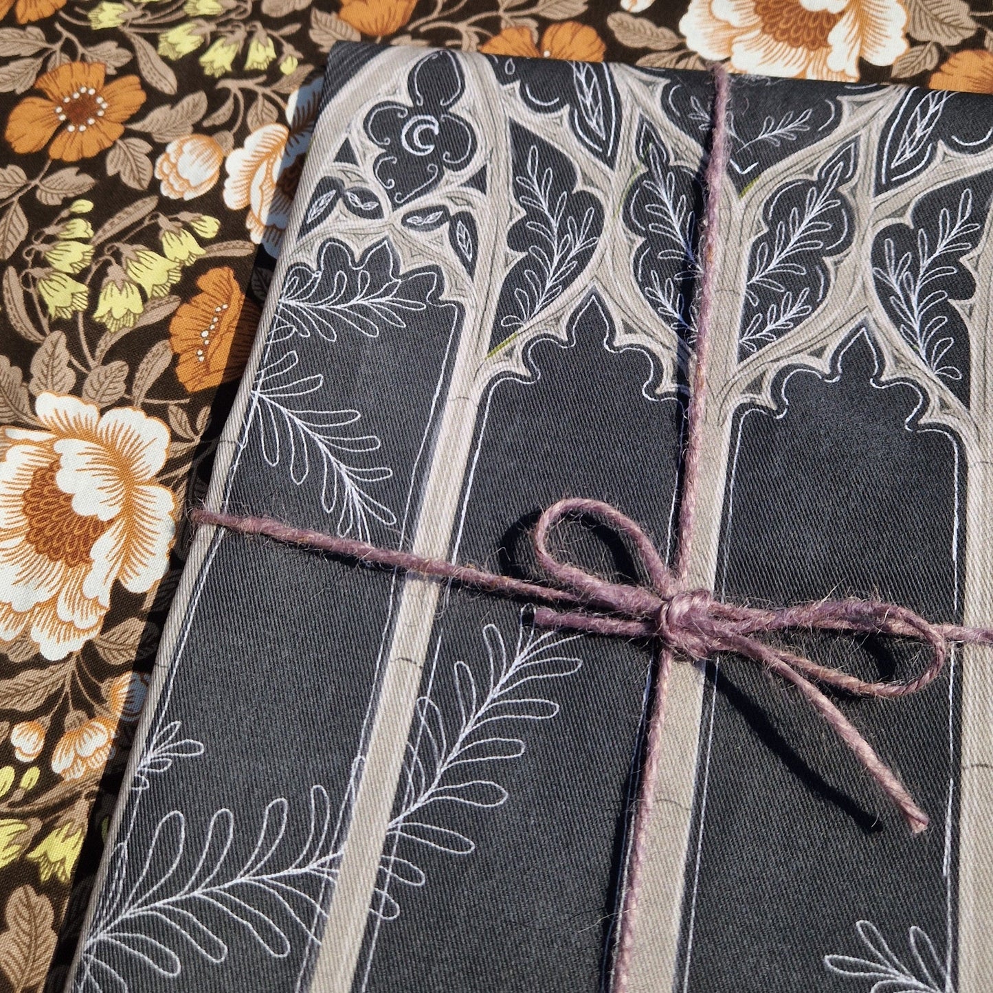 Close up of the folded grey teatowel on a retro floral patterned fabric. The teatowel is wrapped in purple twine and has a light grey and white cathedral window design partially visible.
