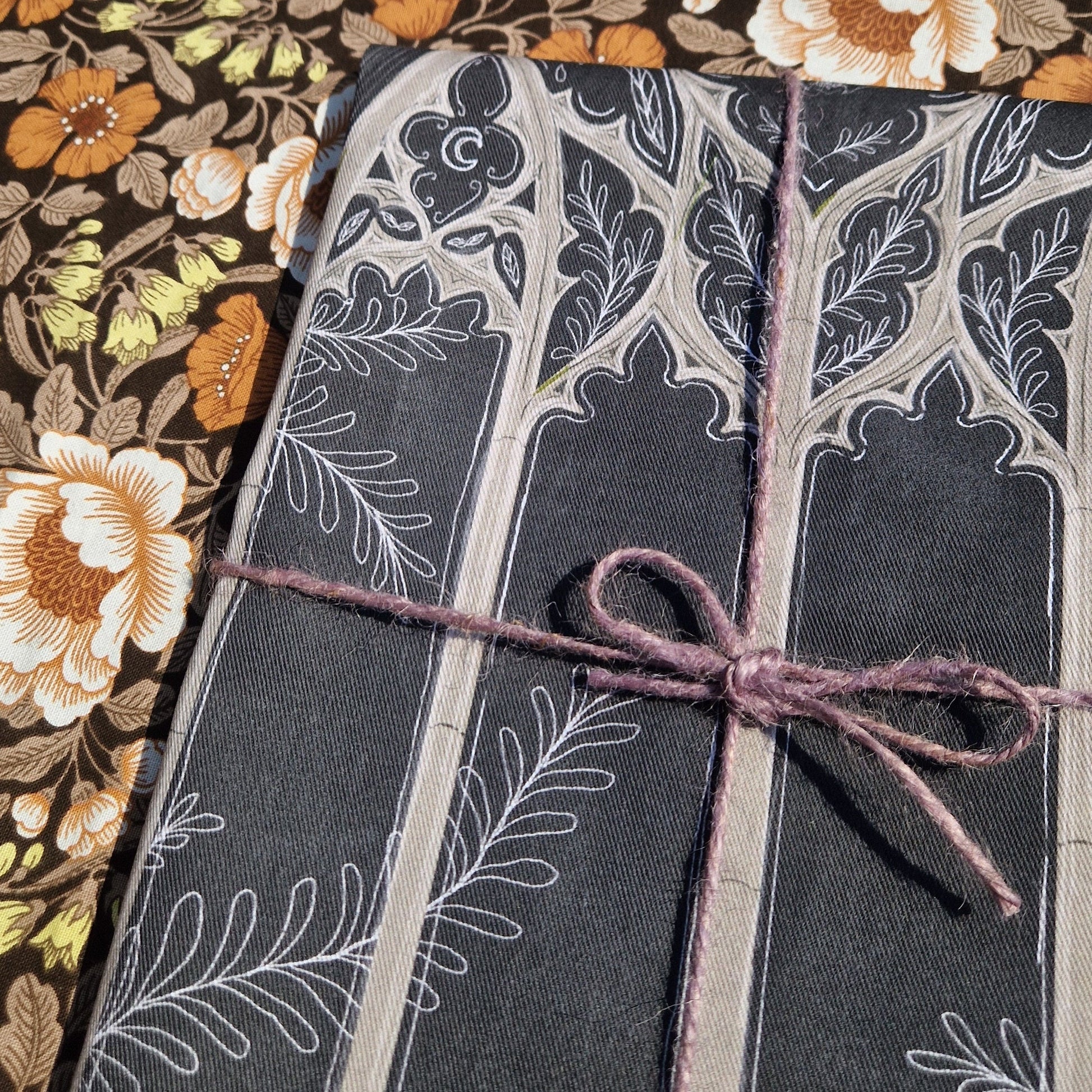 Close up of the folded grey teatowel on a retro floral patterned fabric. The teatowel is wrapped in purple twine and has a light grey and white cathedral window design partially visible.
