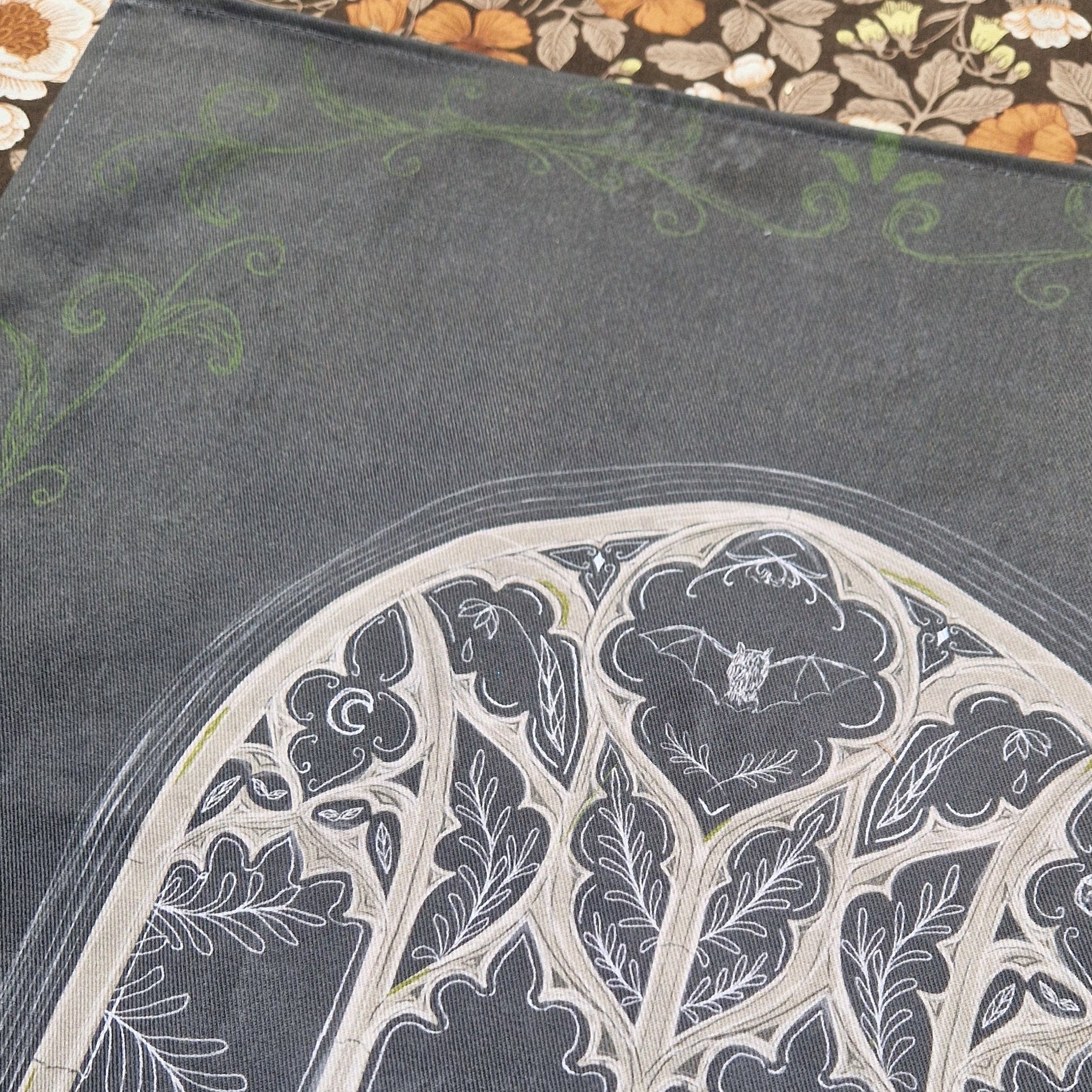 The top of the dark grey teatowel showing the top section of the cathedral window design with the nature inspired window design. Across the sides of the teatowel you can see the green leafy filigree inspired pattern.