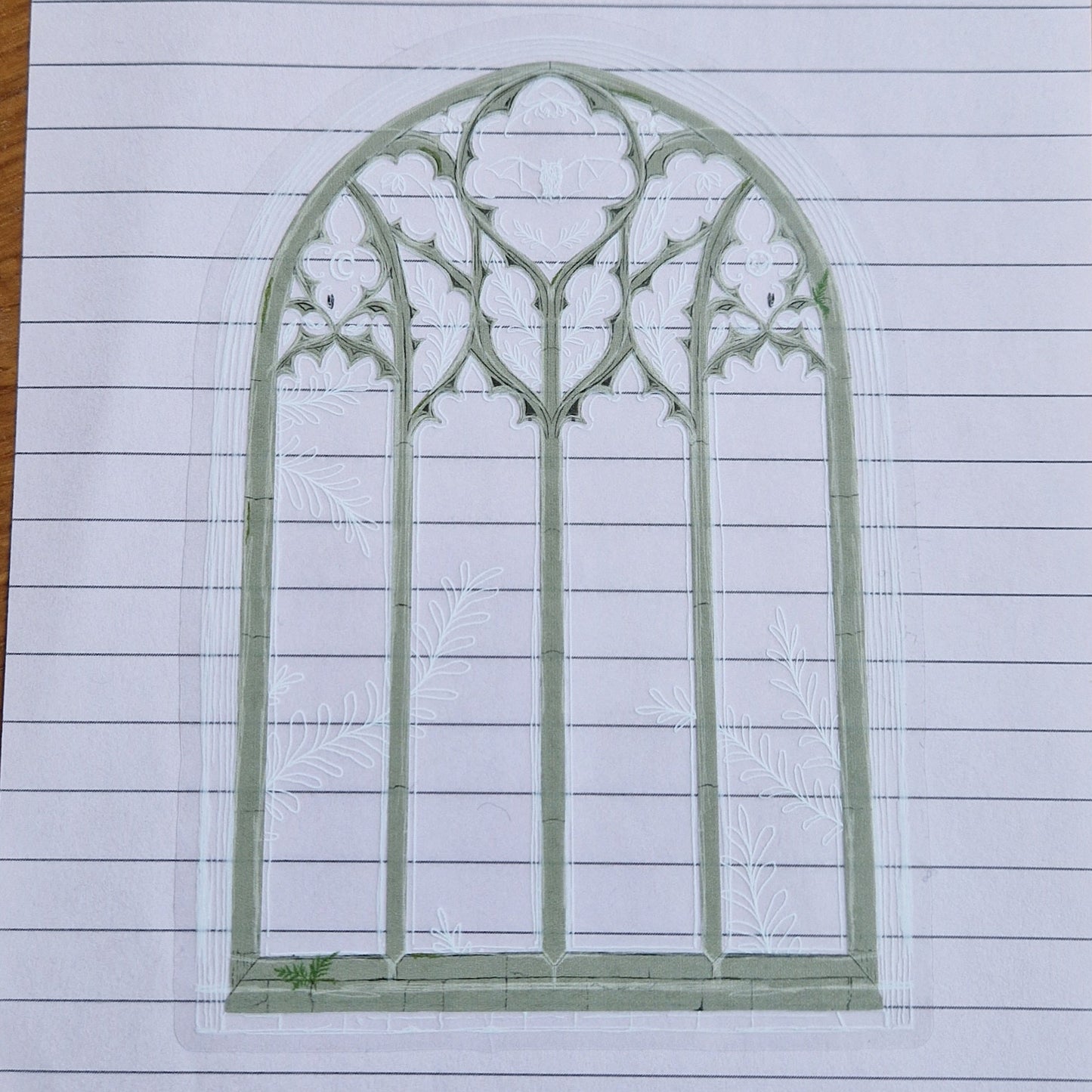 A sticker with a transparent background, in the shape of a gothic cathedral window. The main shape of the window is in a grey brick colour with white sketch lines around                                 the edges and inside the top of the window depicting a bat, leafy fronds, flowers and the mood. Small green patches of moss and ferns grow out of parts of the window.