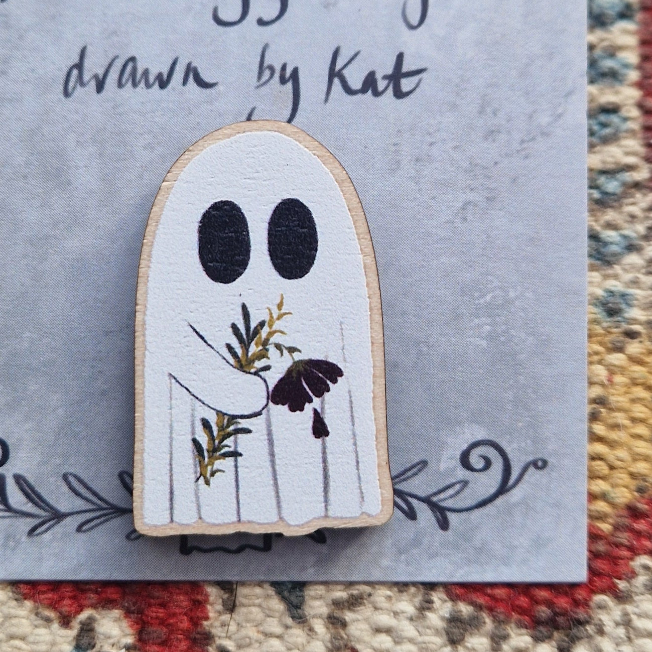 A close up picture of the ghost carrying a deep red flower with a petal falling off pni badge on a grey backing card.