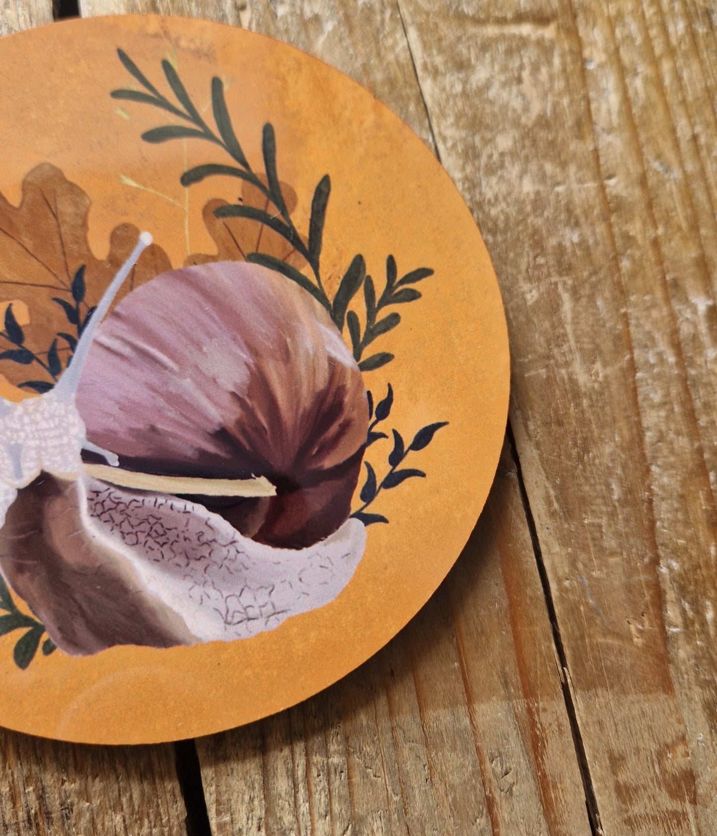 A close up of the round yellow snail coaster showing the detail of the pink and brown garden snail surrounded by dark green leafy vines and warm brown oak leaves. Behind the coaster are wooden planks.