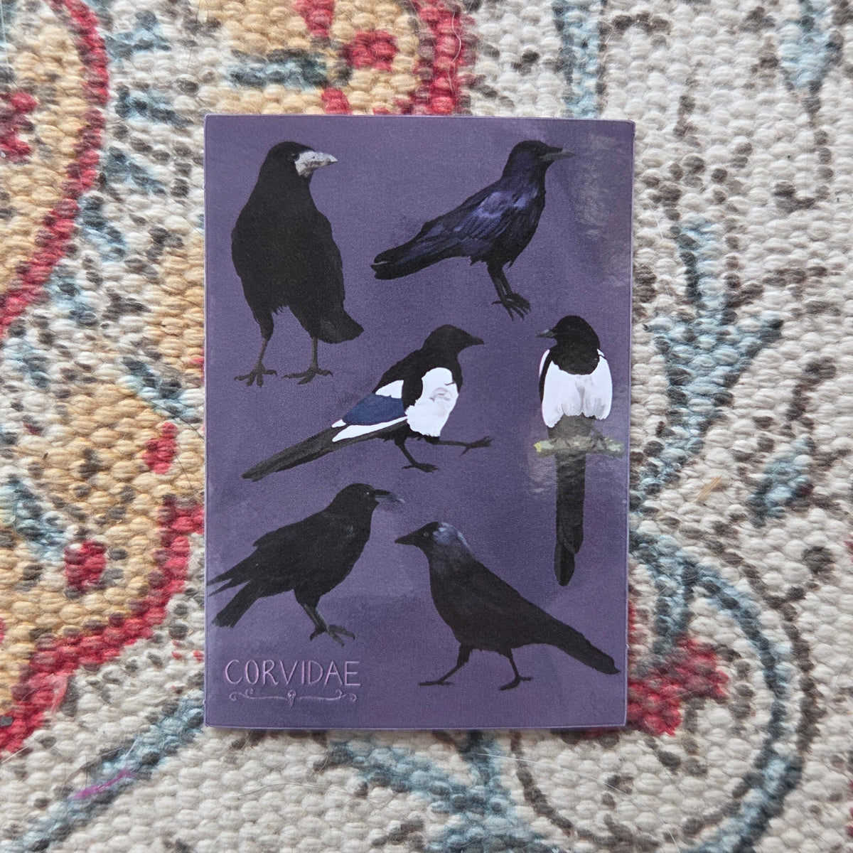A purple rectangular sticker sits on a patterned fabric background. The sticker shows six corvids and the word 'CORVIDAE' in the bottom left corner. 