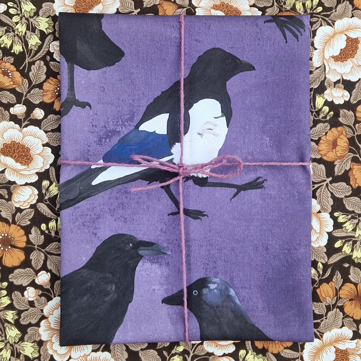 A folded purple teatowel wrapped in a lilac purple twine sits on a floral fabric. The purple teatowel has a magpie visible in the middle and parts of a raven and jackdaw visible on the bottom.
