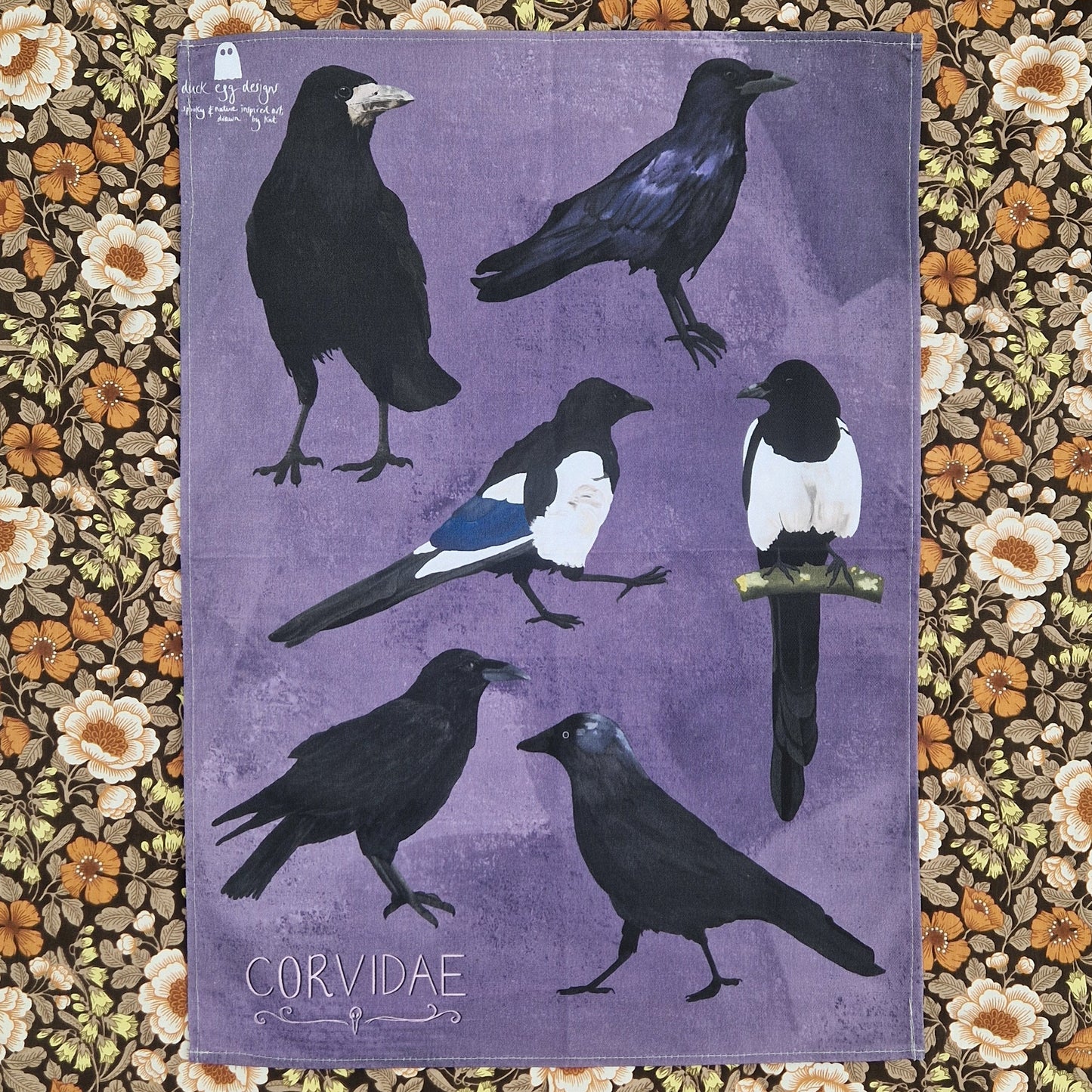 A purple tea towel lies unfolded on top of a floral background. The tea towel features six birds from the corvid family with the word 'CORVIDAE' in light pink writing in the bottom left above a simple raven skull design.