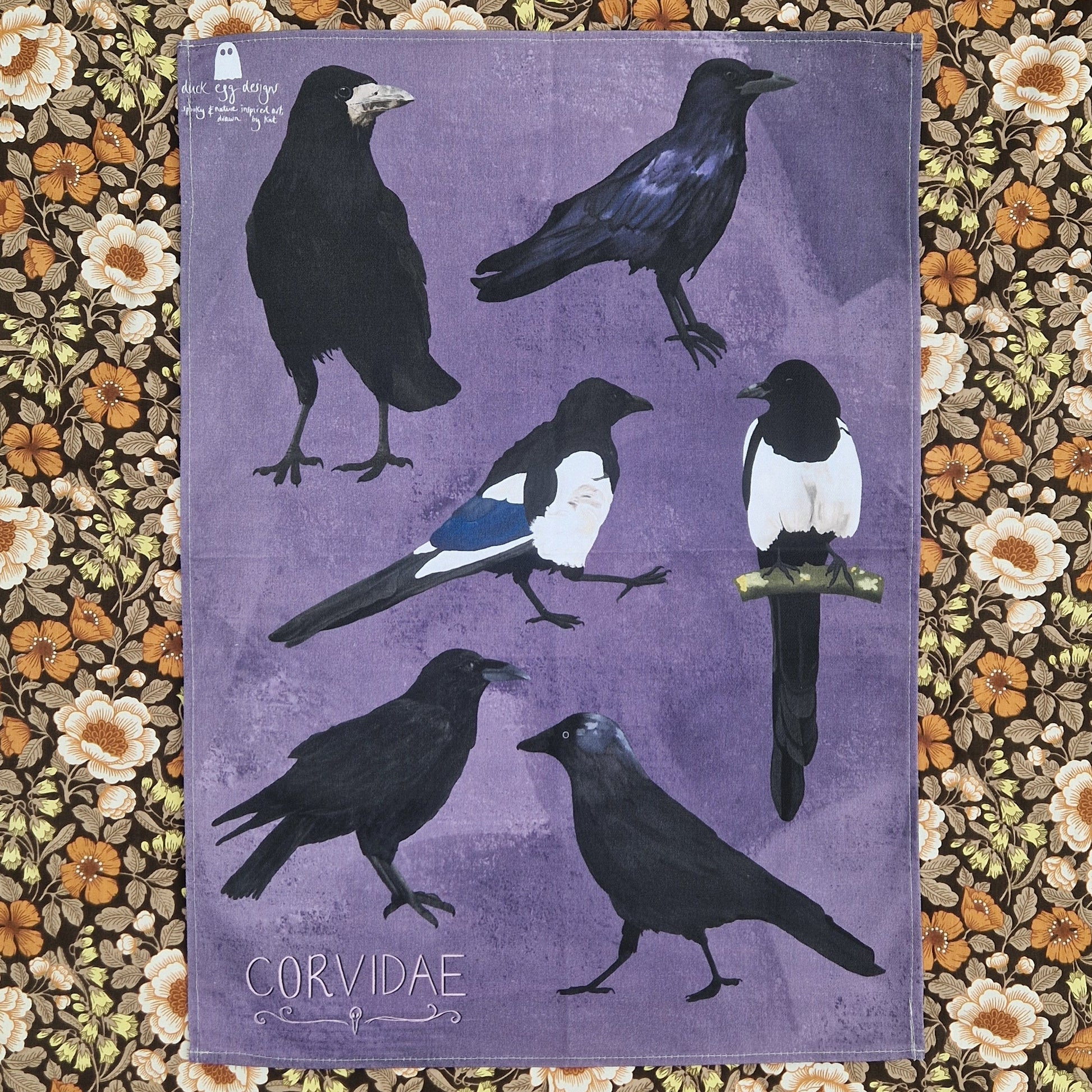 A purple tea towel lies unfolded on top of a floral background. The tea towel features six birds from the corvid family with the word 'CORVIDAE' in light pink writing in the bottom left above a simple raven skull design.