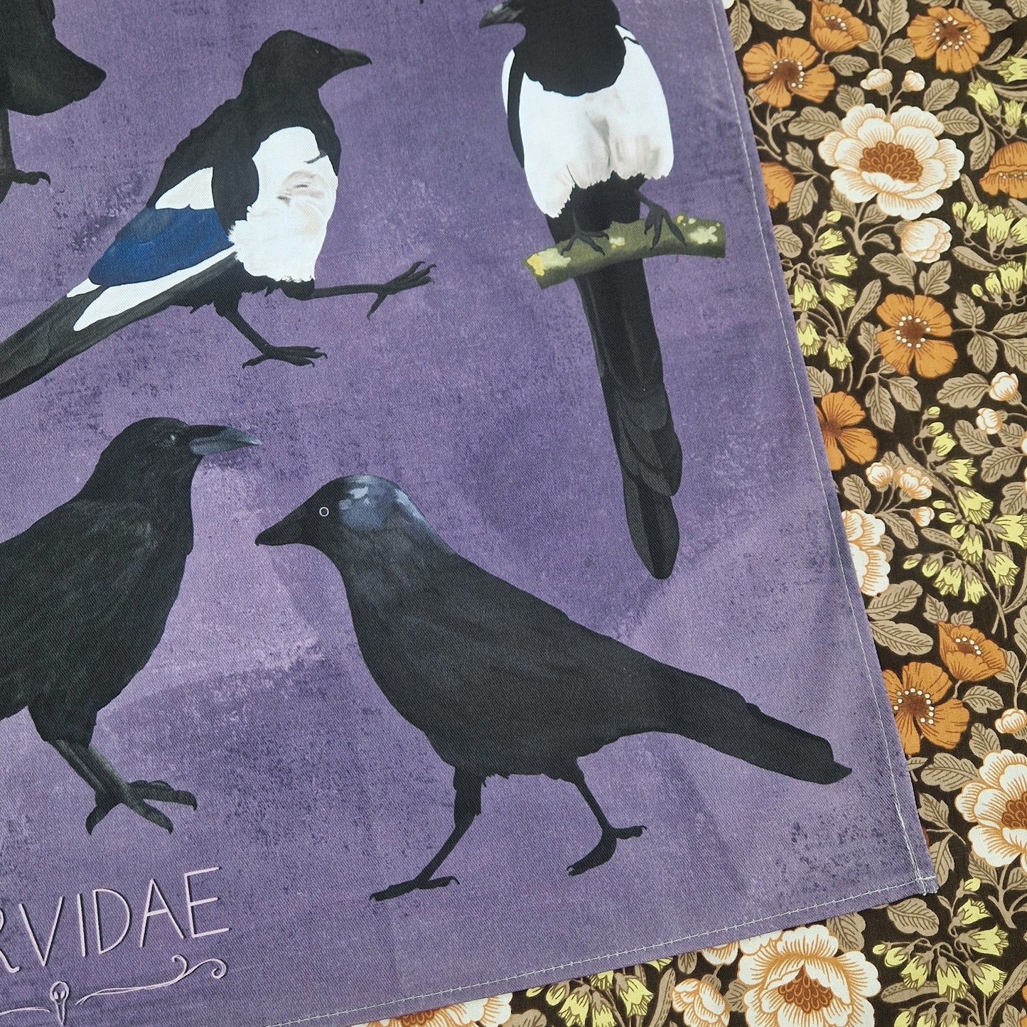 The bottom right of the purple teatowel displaying the jackdaw with the crow to the left of them and two magpies above. To the right of the tea towel you can see a floral patterned background.