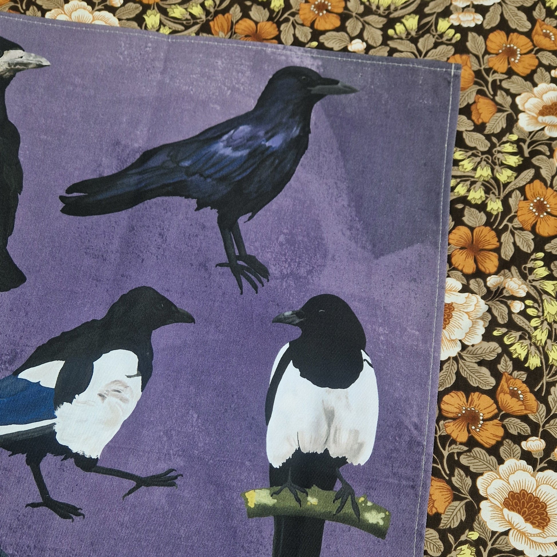 The top right of the teatowel showing the detail in the crow illustration as well as part of the rook to the left of it and the two magpies below. To the right of the purple tea towel you can see a floral patterned background.
