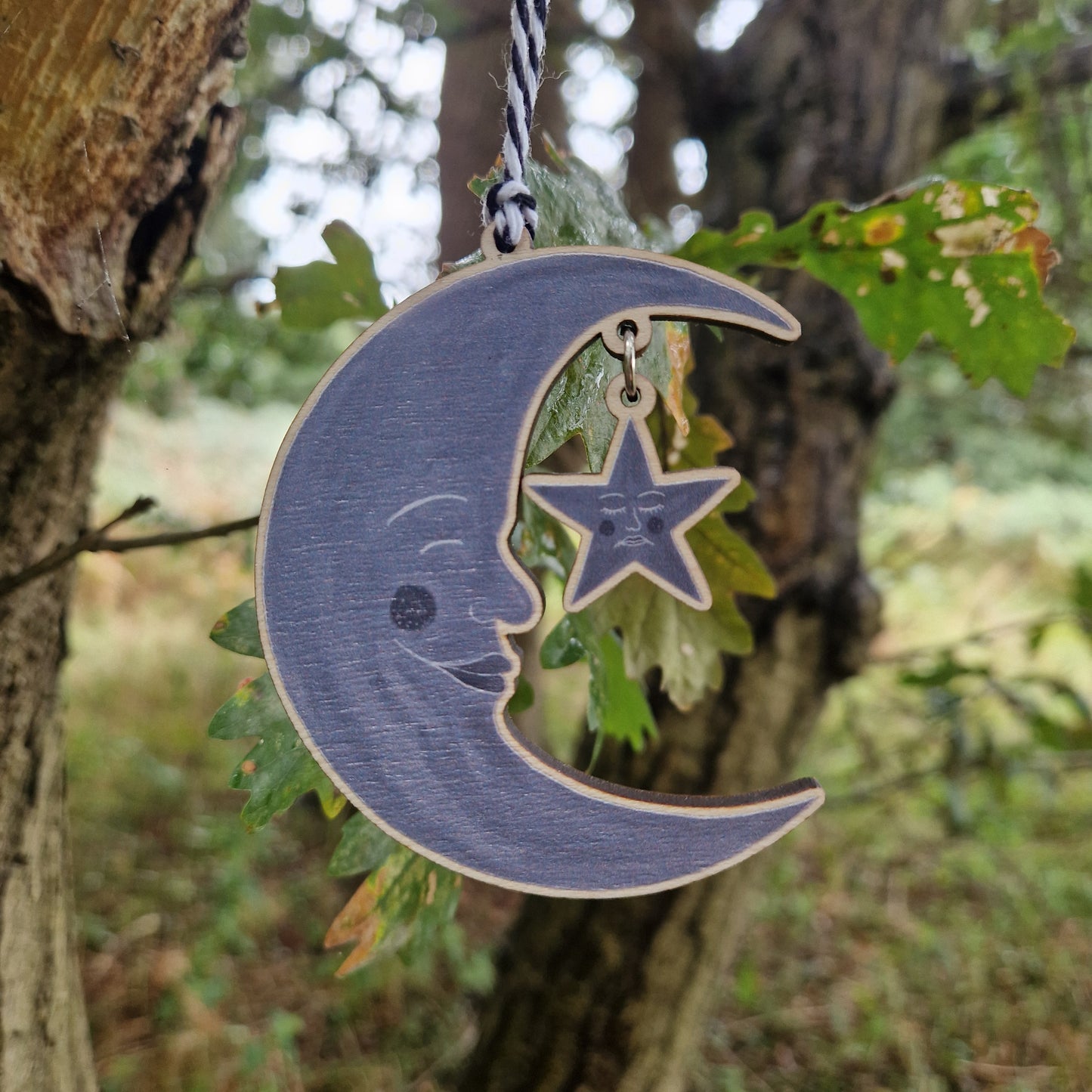 A wooden crescent moon with a star hanging from it on a silver jump ring hangs from a black and white bakers twine on a tree. 