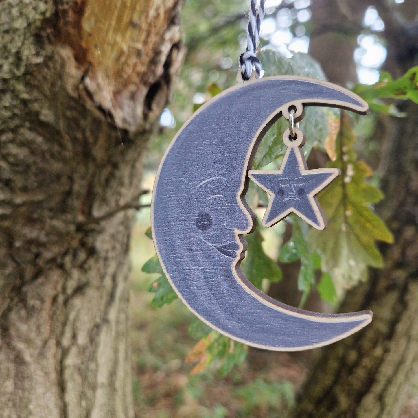 A wooden grey crescent moon with a grey star hanging from it by a silver coloured jump ring hangs next to a tree in front of some green leaves.