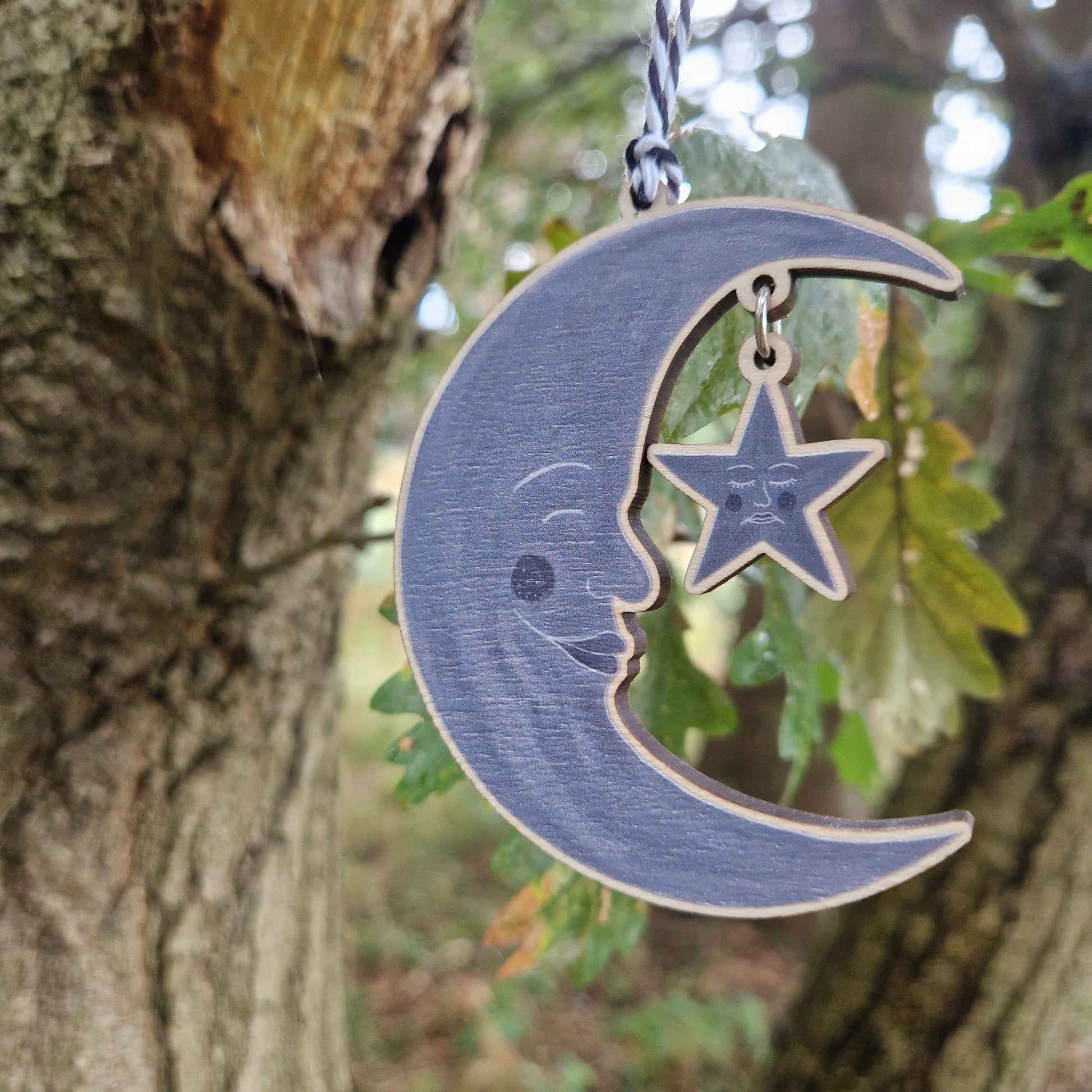 A wooden grey crescent moon with a grey star hanging from it by a silver coloured jump ring hangs next to a tree in front of some green leaves.