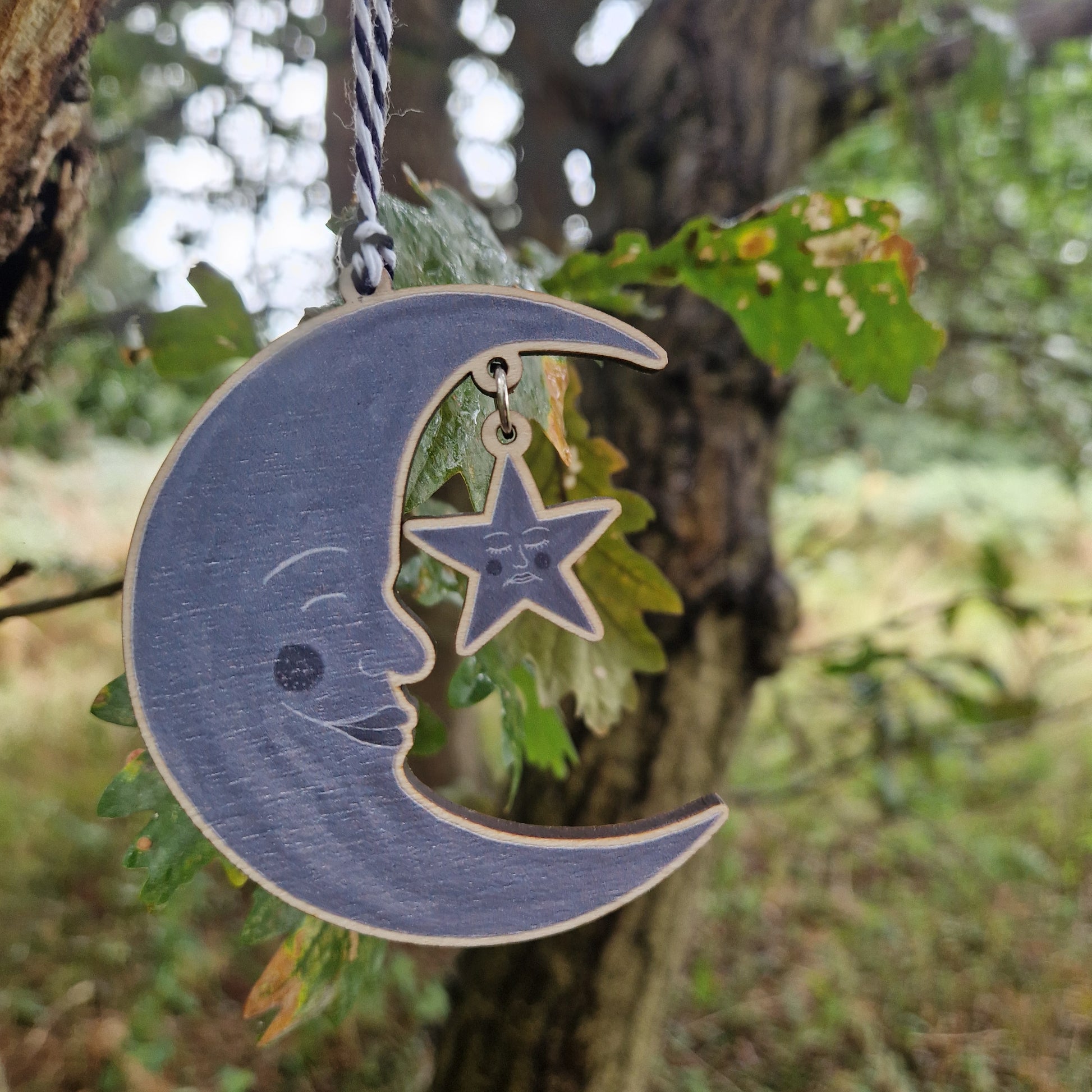 A wooden grey crescent moon with a grey start hanging from it by a silver coloured jump ring hangs next to a tree in front of some green leaves.