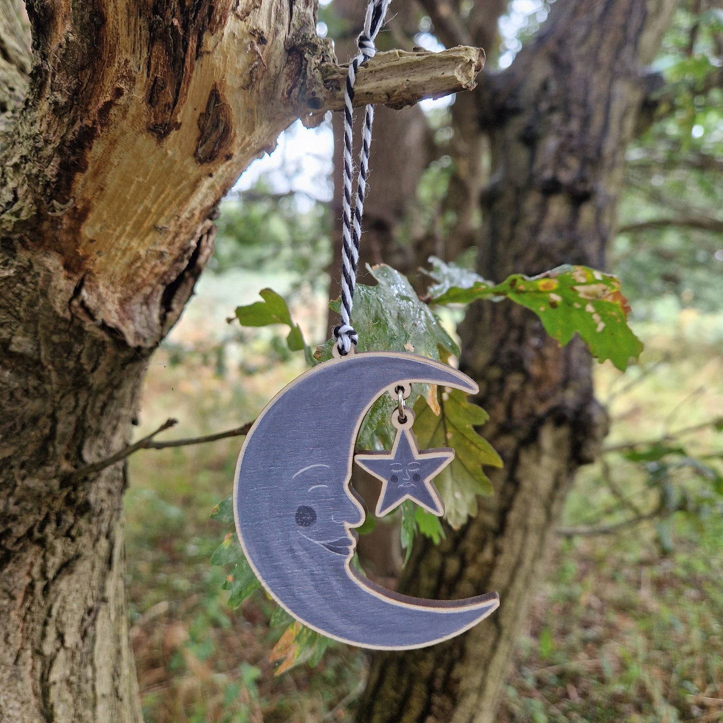 A wooden grey crescent moon with a grey start hanging from it by a silver coloured jump ring hangs from a tree in front of some green leaves by a black and white bakers twine.