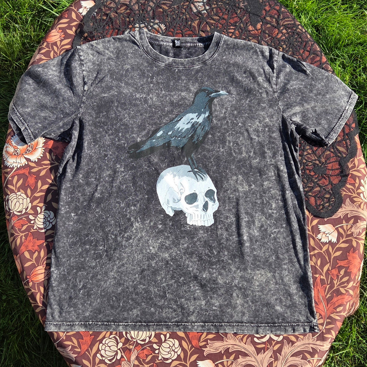 A acidwash black t-shirt lies on a brown floral patterned fabric with a black lace overlay. The shirt features a screenprinted design of a crow stood atop of a human skull.