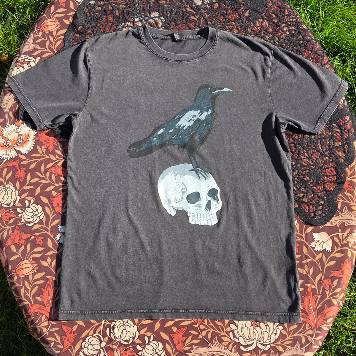A stonewash black t-shirt lies on a brown floral patterned fabric with a black lace overlay. The shirt features a screenprinted design of a crow stood atop of a human skull.