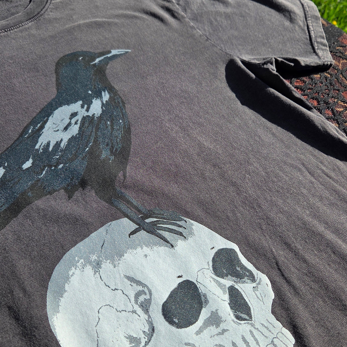 A close up of a stonewash black t-shirt laying on a brown floral patterned fabric with a black lace overlay. The shirt features a screenprinted design of a crow stood atop of a human skull.