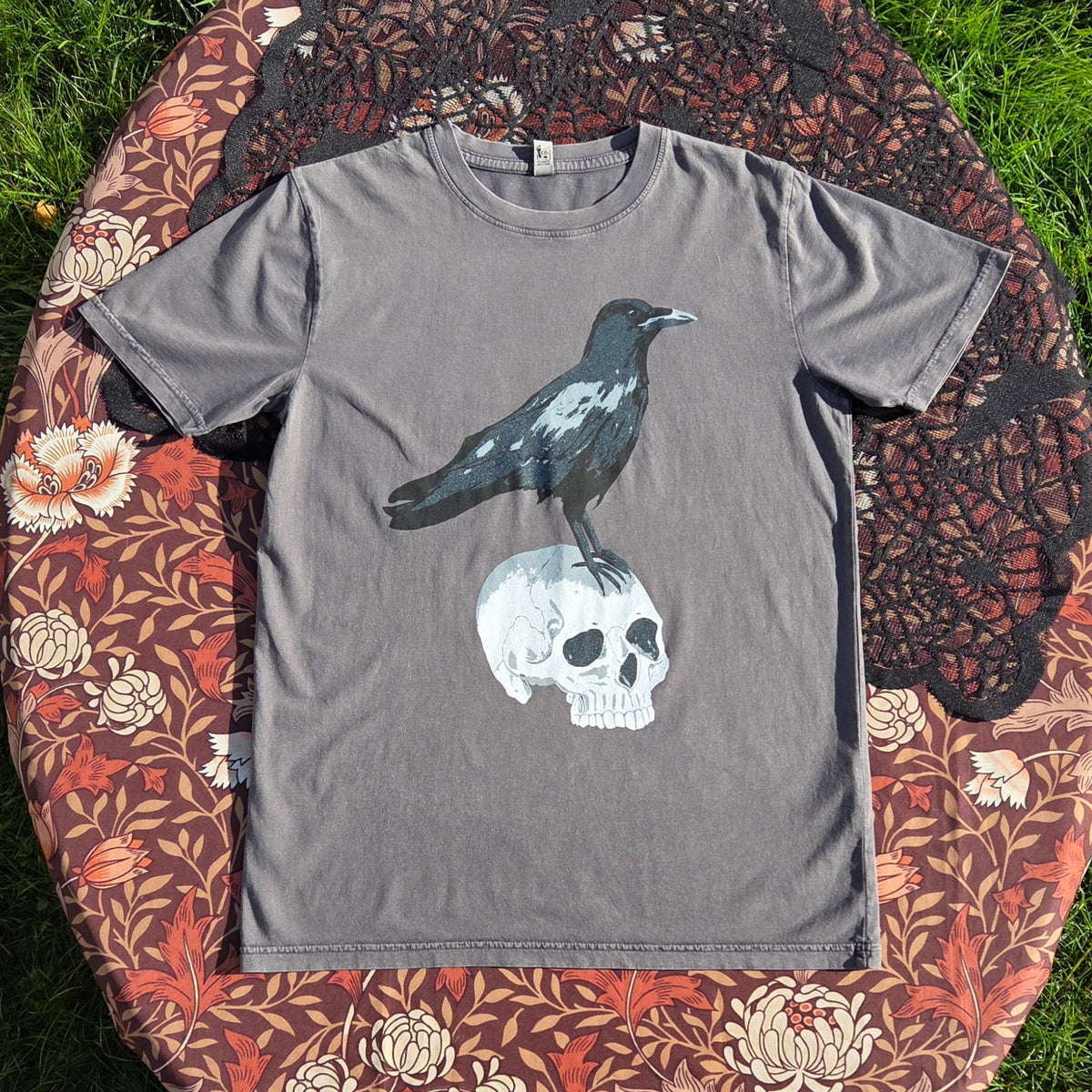 A stonewash grey t-shirt lies on a brown floral patterned fabric with a black lace overlay. The shirt features a screenprinted design of a crow stood atop of a human skull.