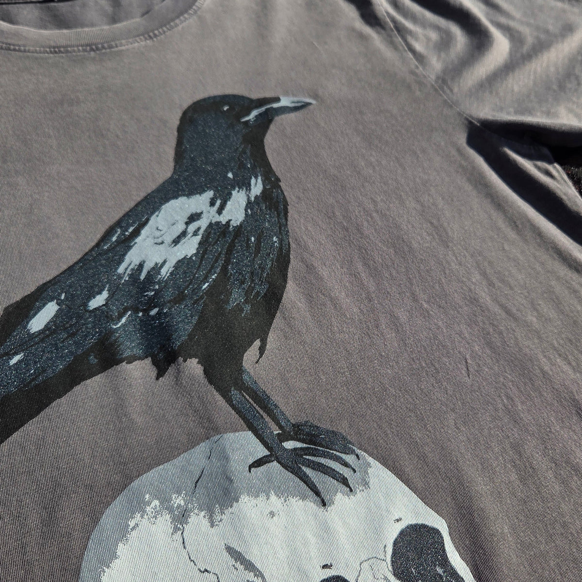 A close up of a stonewash grey t-shirt laying on a brown floral patterned fabric with a black lace overlay. The shirt features a screenprinted design of a crow stood atop of a human skull.