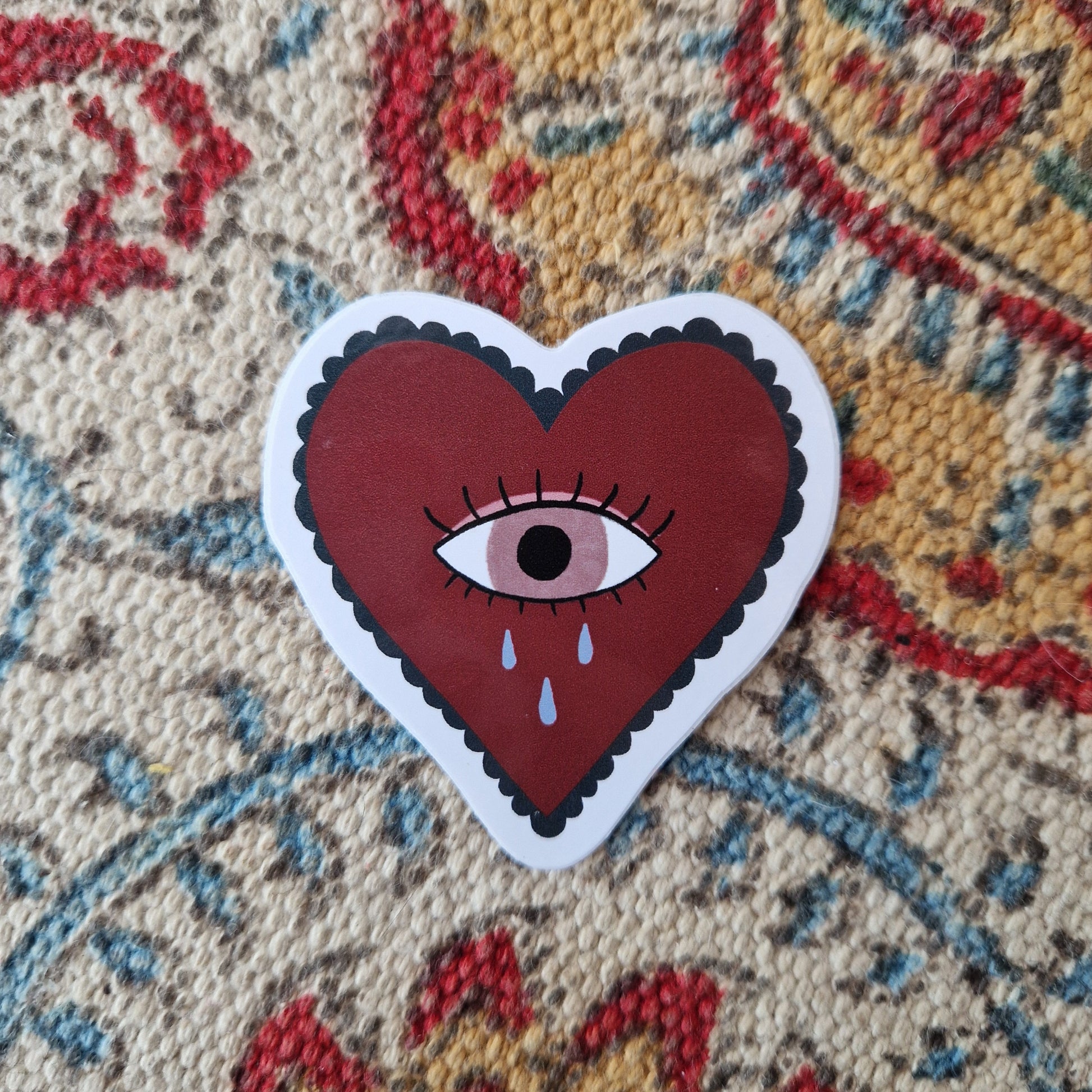 Rich red heart sticker with an eye in the middle with three tear drops falling from it. Behind the sticker is a patterned white, yellow, blue and red fabric.